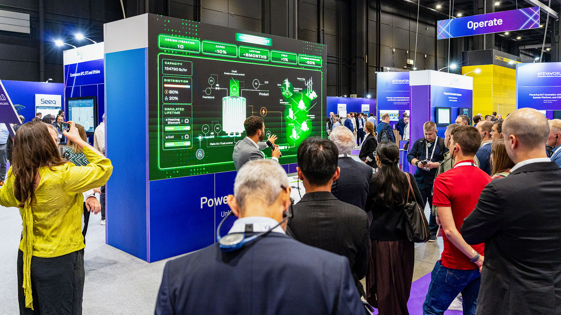 A crowd of people gathers at a technology trade show, watching a presenter explain information displayed on a large, futuristic digital screen about power management. Attendees listen and take photos in a busy, modern exhibition hall.