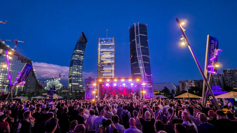 A large crowd attends an outdoor concert at night in a city, with colorful stage lights and tall, modern skyscrapers lit up in the background. People are taking photos and enjoying the lively atmosphere.