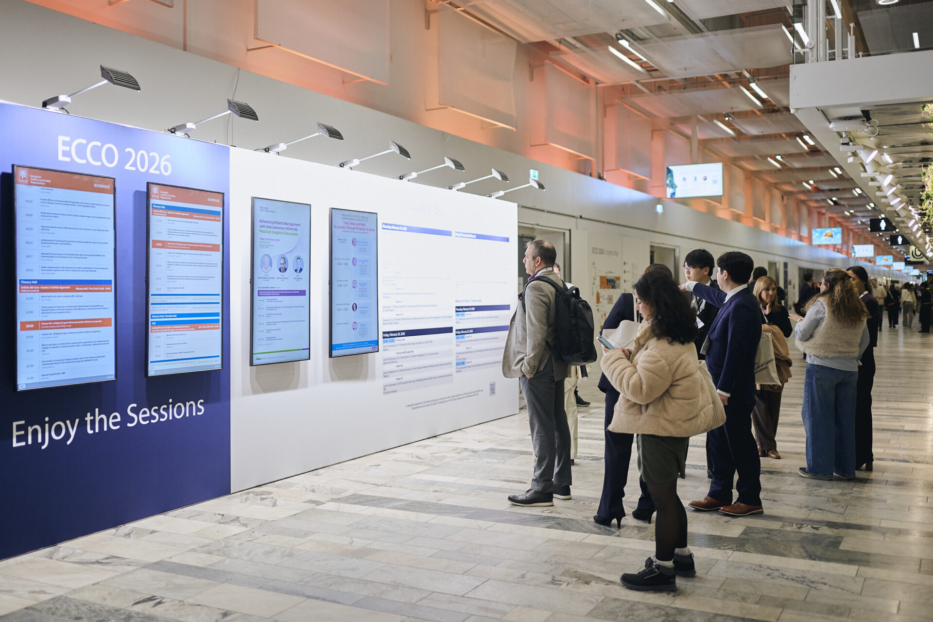 A group of people stands in front of large digital screens displaying conference schedules and information at an indoor event called ECCO 2026. The setting is modern and well-lit.