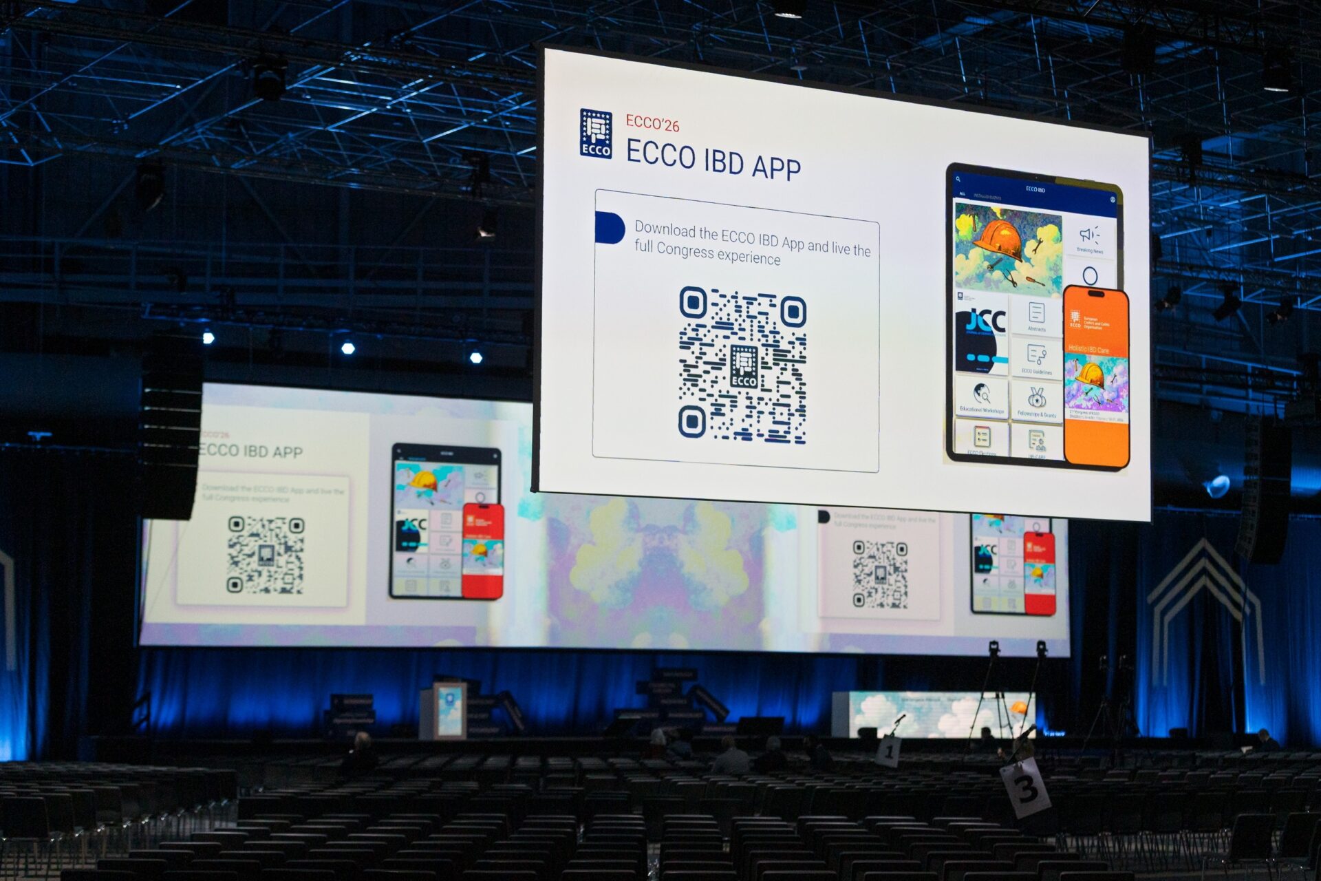 A large screen at a conference displays information about the ECCO IBD app, including QR codes and images of the app’s interface. The auditorium below is filled with empty chairs and blue lighting.