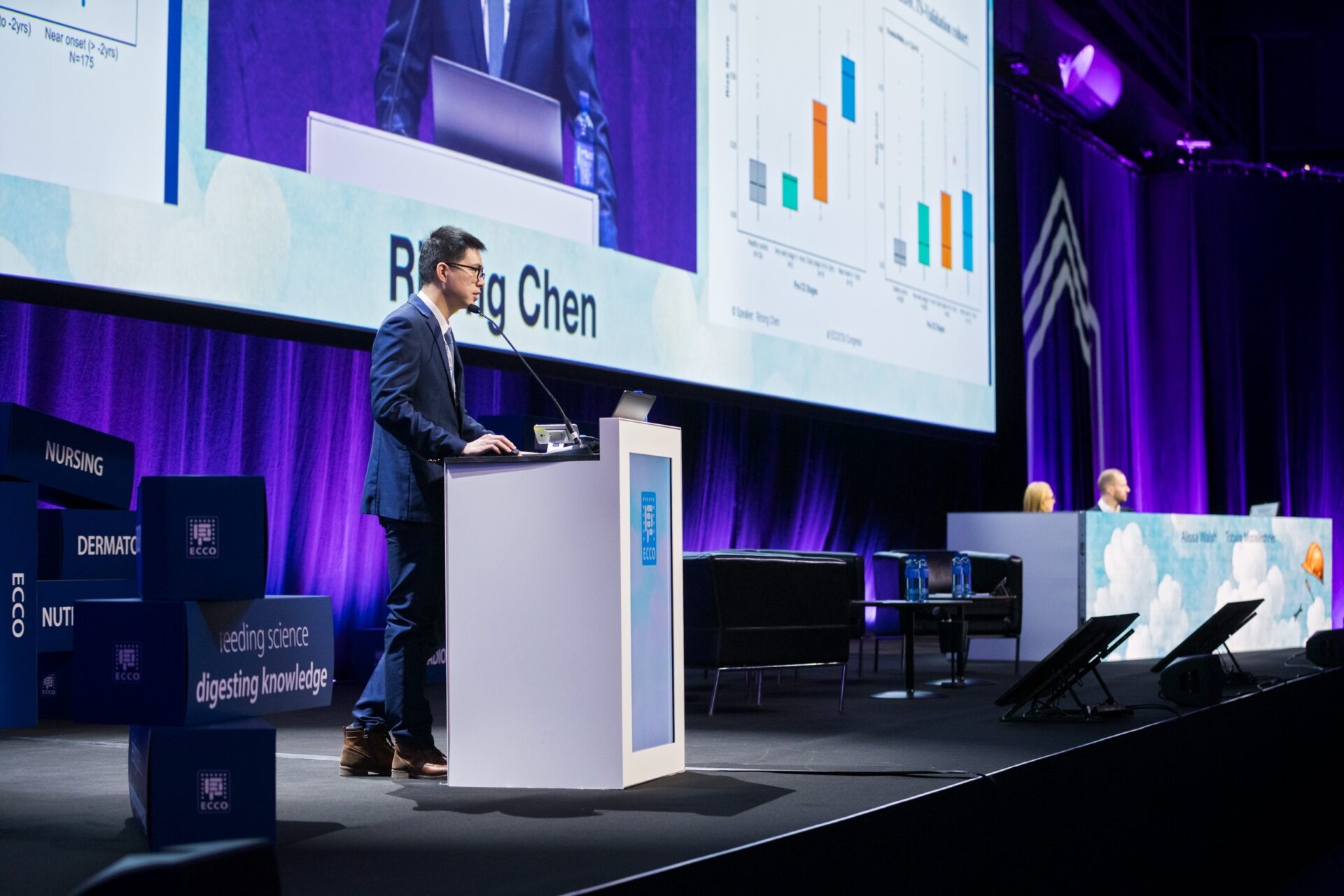 A man in a suit is speaking at a podium on stage, with charts displayed on a large screen behind him. Two people are seated at a panel table nearby. The lighting is purple and blue, creating a professional conference atmosphere.