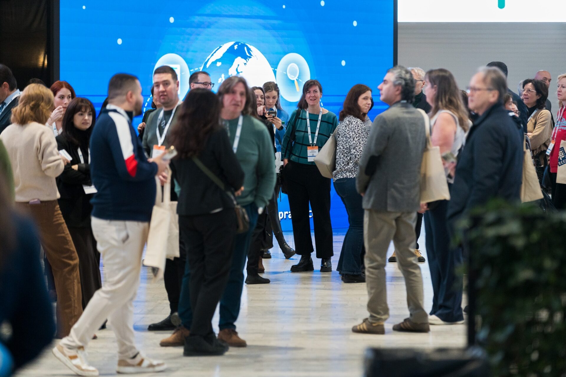 A group of people are chatting and mingling at an indoor event or conference, with a large blue screen displaying a globe and icons in the background. Most attendees are wearing name tags and carrying tote bags.