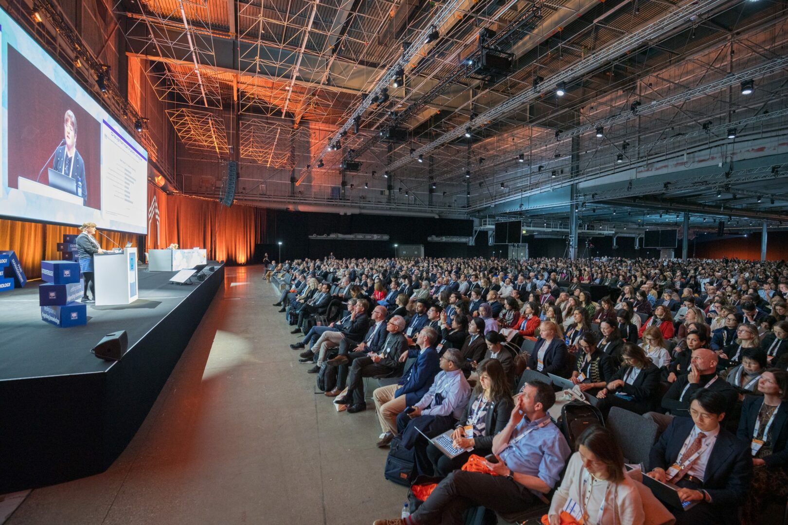A large audience sits in rows, listening intently to a speaker presenting on stage at a well-lit conference held in a spacious industrial-style venue.