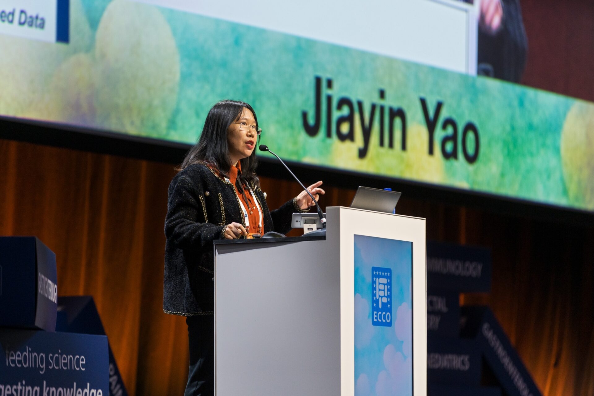 A woman stands at a podium giving a presentation, with the name “Jiayin Yao” displayed on a large screen behind her at a conference.