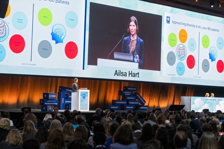 A woman stands at a podium speaking to a large audience at a conference, with colorful medical diagrams and her name, Ailsa Hart, displayed on large screens behind her.