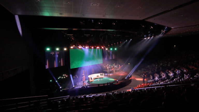 A brightly lit stage with colorful spotlights and a large screen is set up in a dark auditorium filled with seated audience members watching the performance.