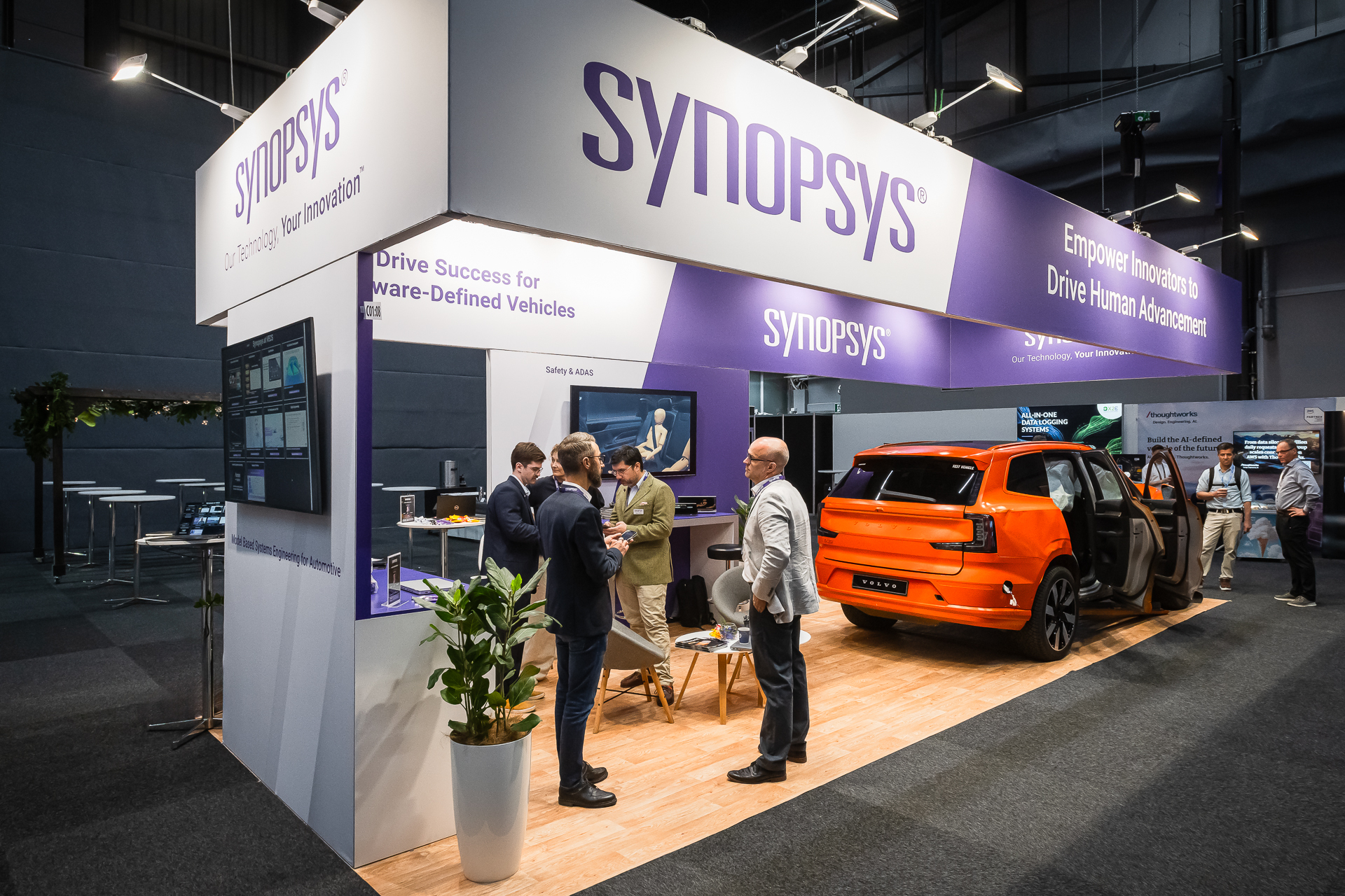 A Synopsys exhibition booth features several people conversing near displays and a bright orange car. The booth displays technology-related signs and exhibits, with “SYNOPSYS” branding visible overhead.