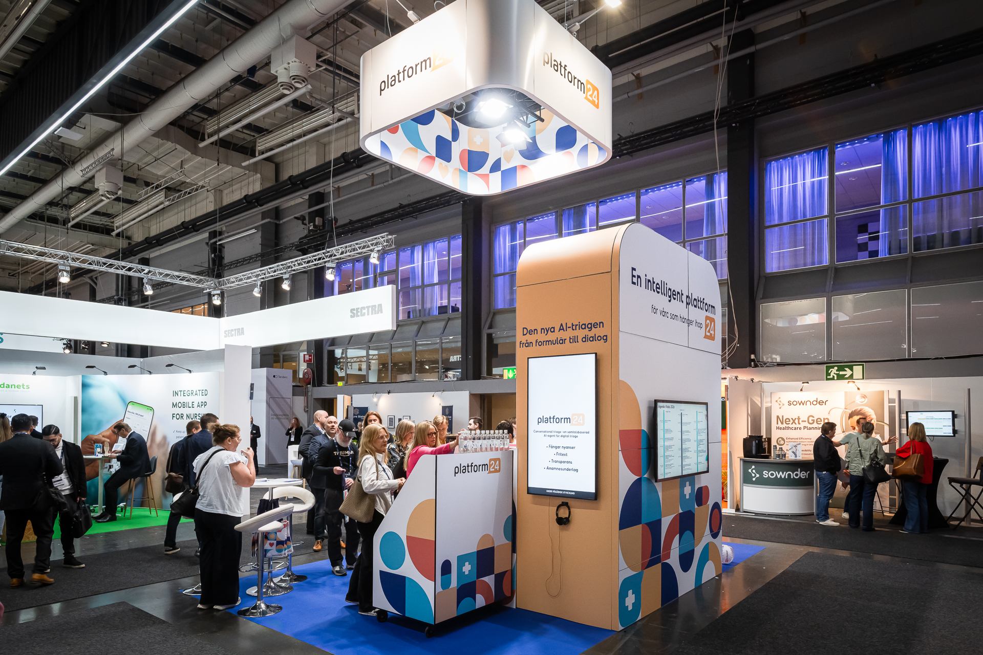 A busy exhibition hall with people networking around modern booths, including a prominent “platform24” stand featuring branded signage, screens, and spotlights under high ceilings.