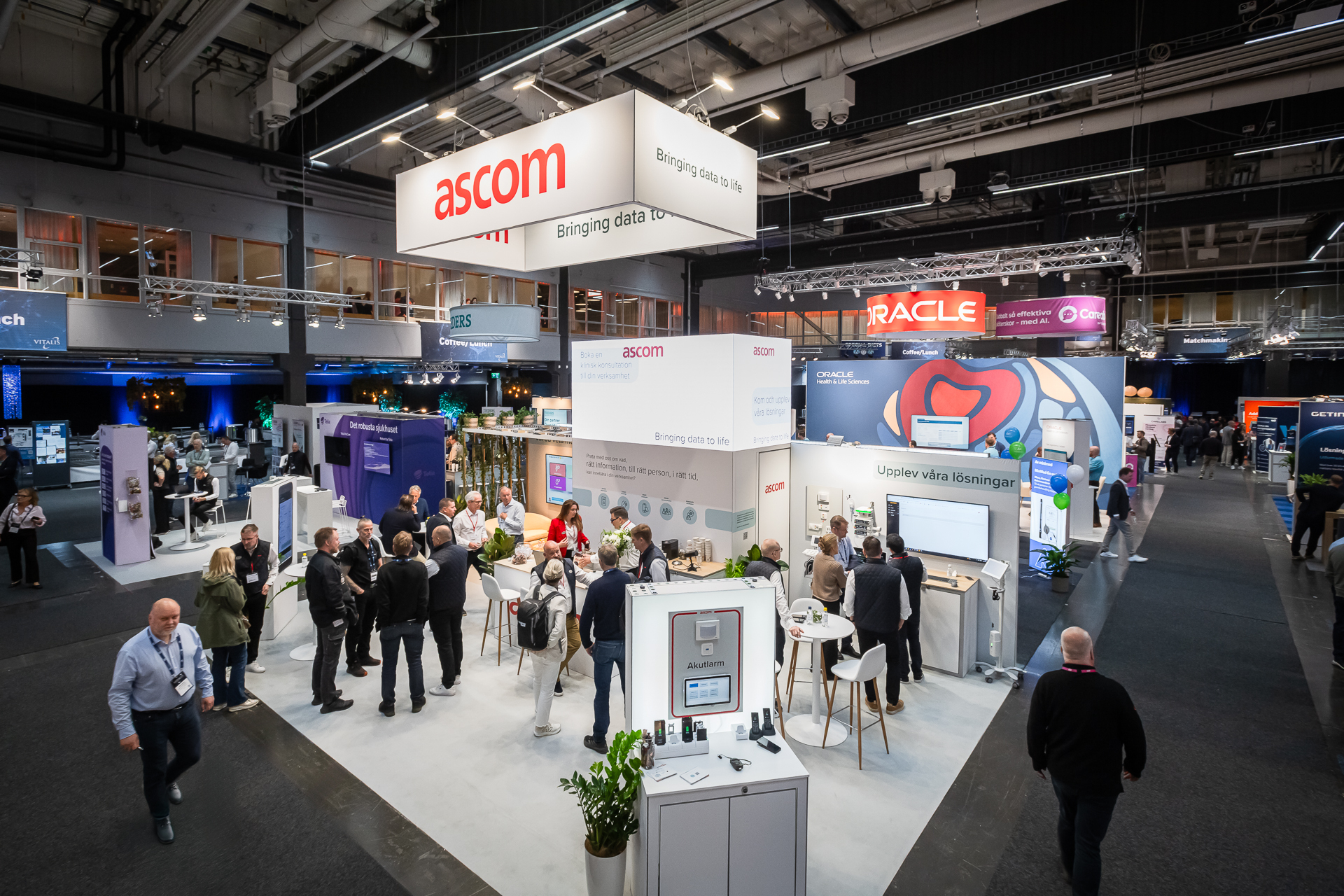 A busy exhibition hall with people visiting various booths, including a prominent white booth with ascom signage and other stands like Oracle nearby; attendees are talking, exploring displays, and networking.