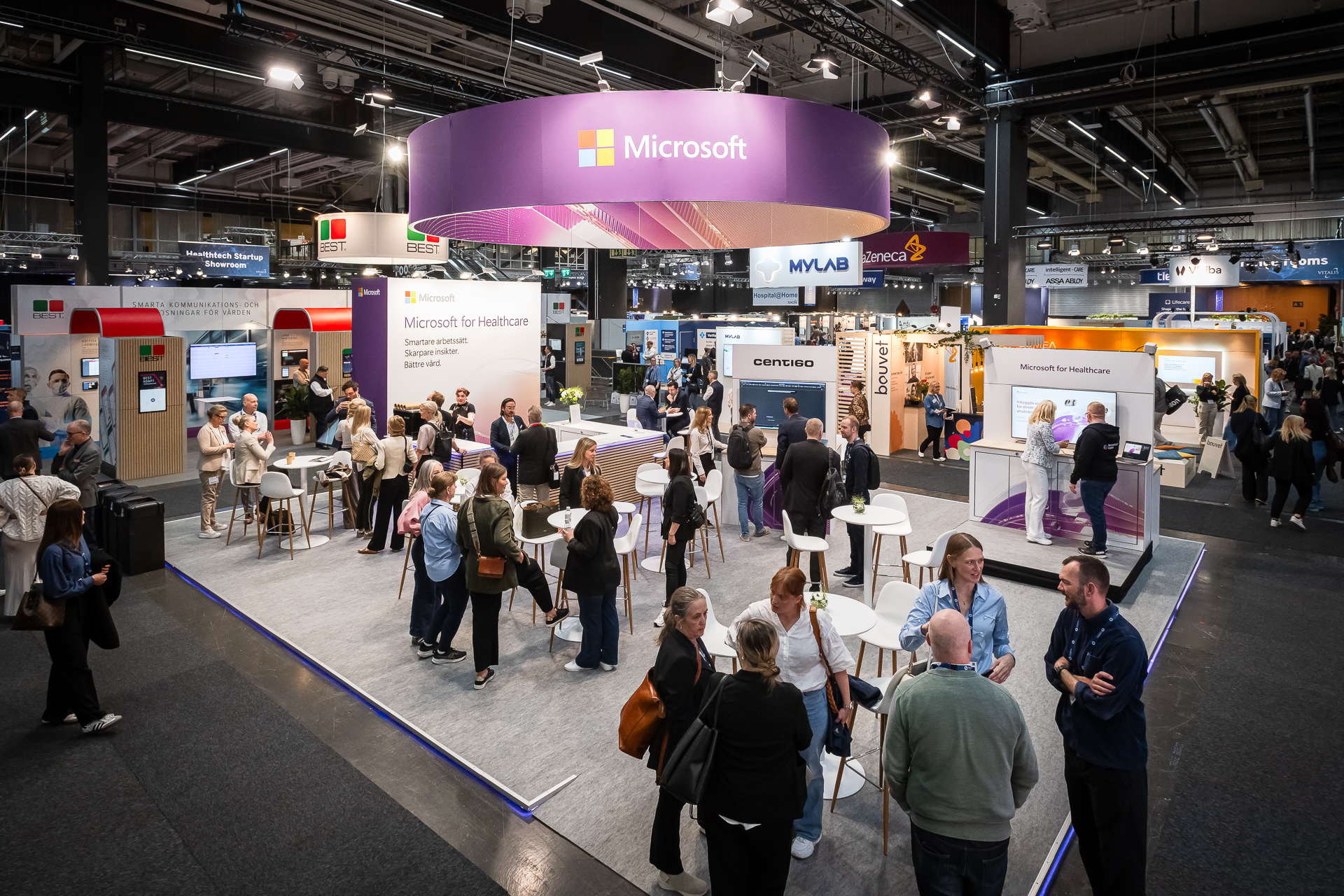 A busy technology conference hall features a large Microsoft booth with people networking, discussing, and exploring various healthcare technology exhibits under bright lights and branded banners.