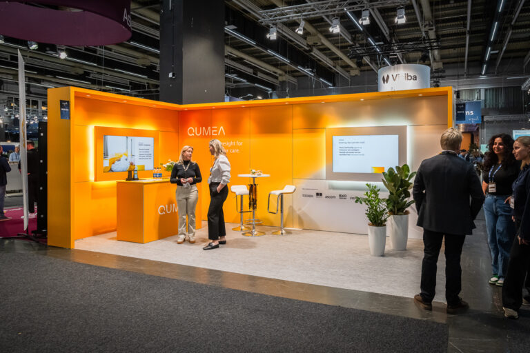 A brightly lit orange exhibition booth with the name QUMEA has two women standing and talking near a counter, with stools, plants, and a screen in the background at a trade show. People are seen walking nearby.