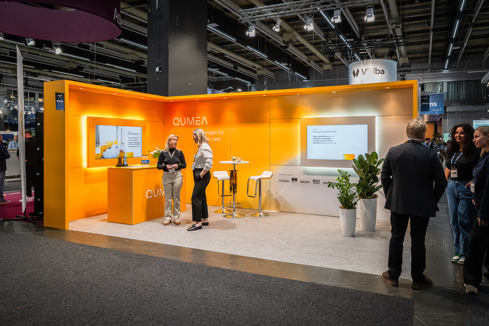 A brightly lit orange exhibition booth with the name QUMEA has two women standing and talking near a counter, with stools, plants, and a screen in the background at a trade show. People are seen walking nearby.