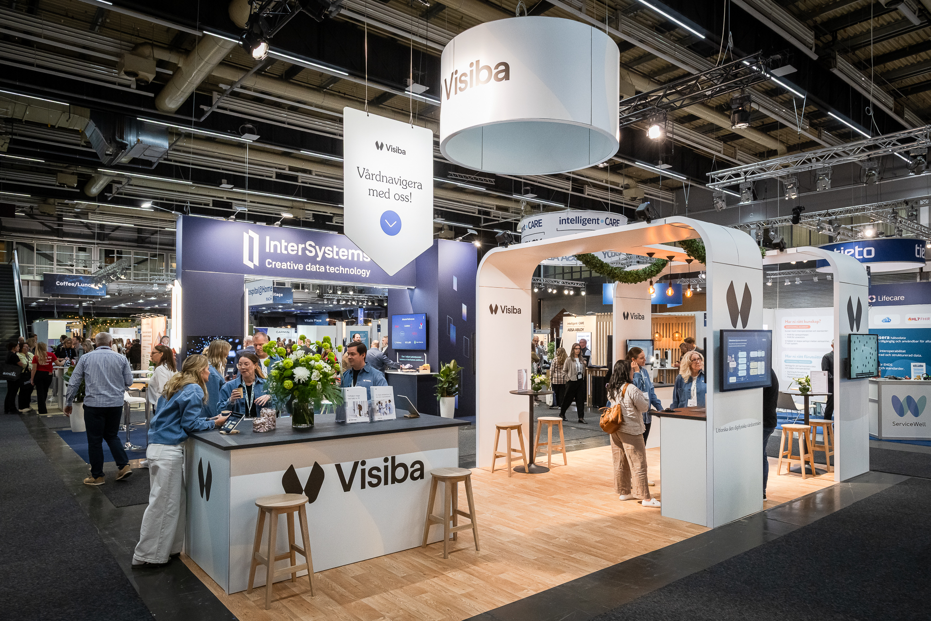 A modern exhibition booth for Visiba at a trade show, with staff interacting with visitors, digital displays, floral arrangements, and branded banners. Other technology company booths are visible in the background.