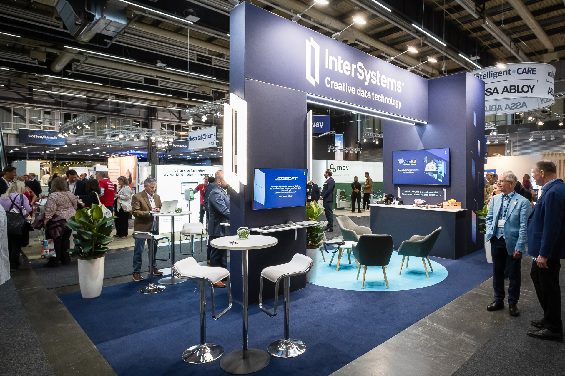 A busy trade show floor features the InterSystems booth with blue carpeting, branded signs, digital screens, tables, chairs, potted plants, and people networking and walking around under bright lights.