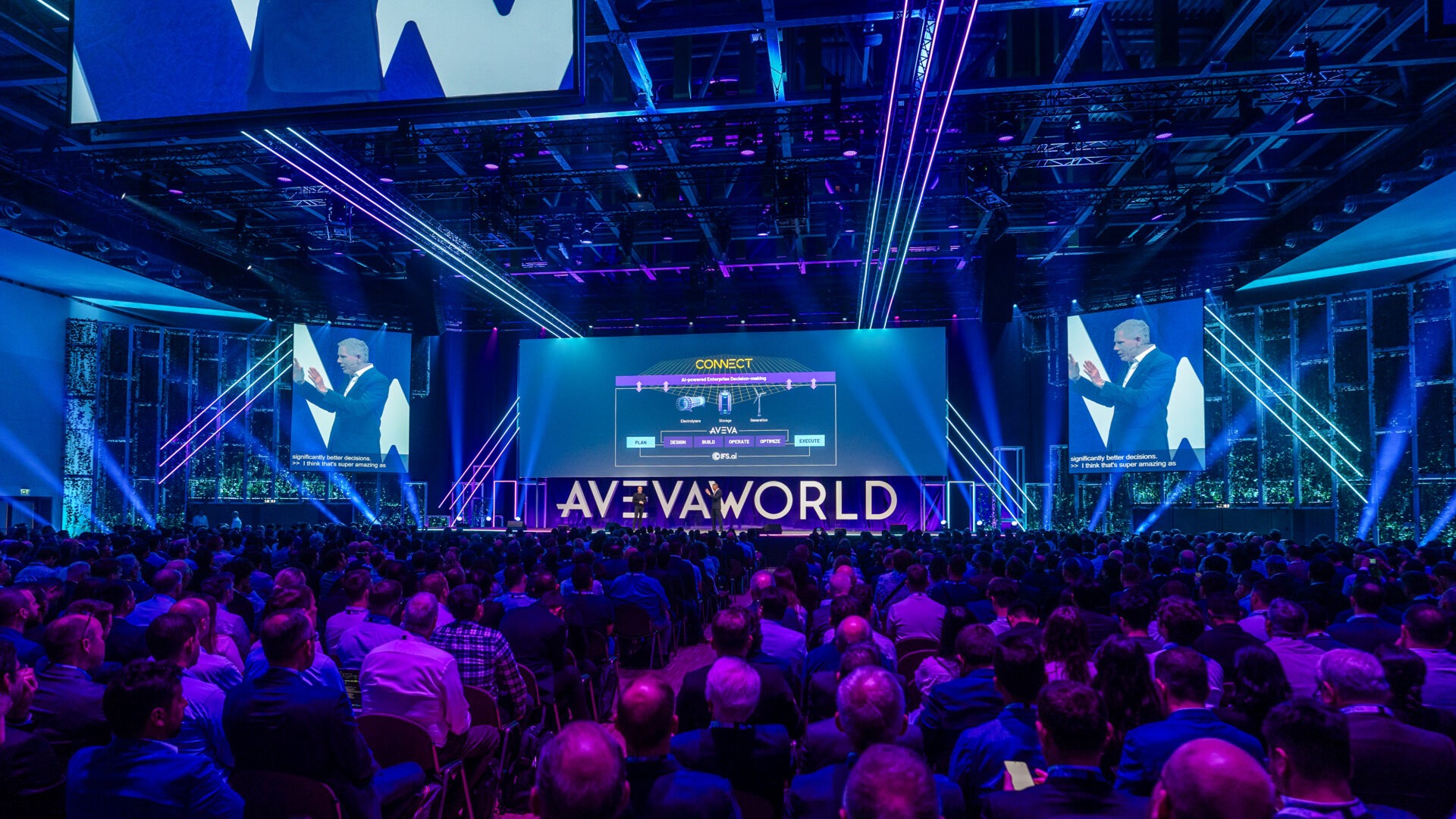 A large crowd attends a tech conference in a spacious, illuminated auditorium. A speaker presents on stage beneath the sign AVEVA WORLD, with presentation slides and live video shown on big screens.