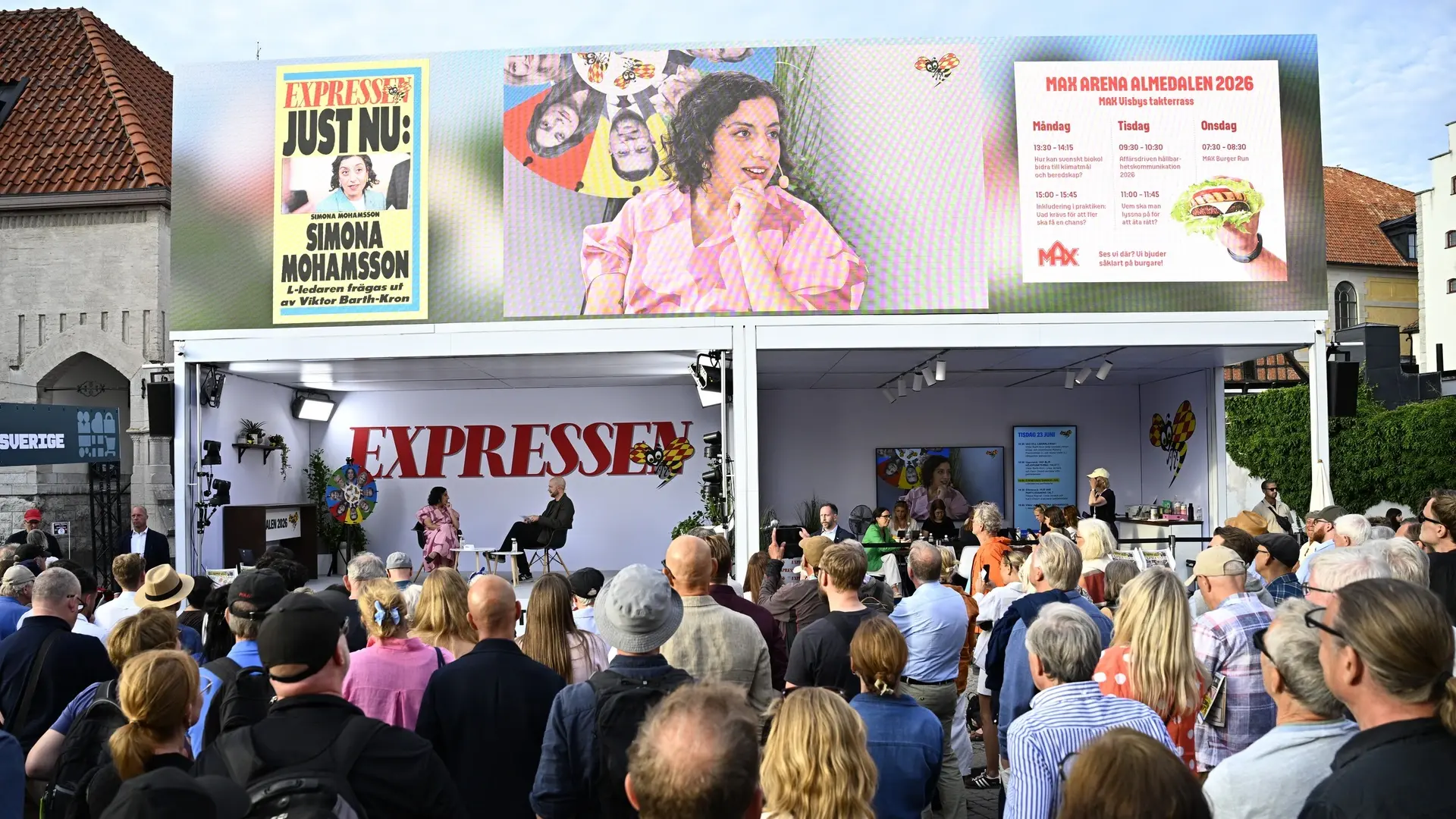 A crowd gathers in front of an outdoor stage with EXPRESSEN branding. Two people speak on stage, while a large digital screen above displays a womans portrait and event information.
