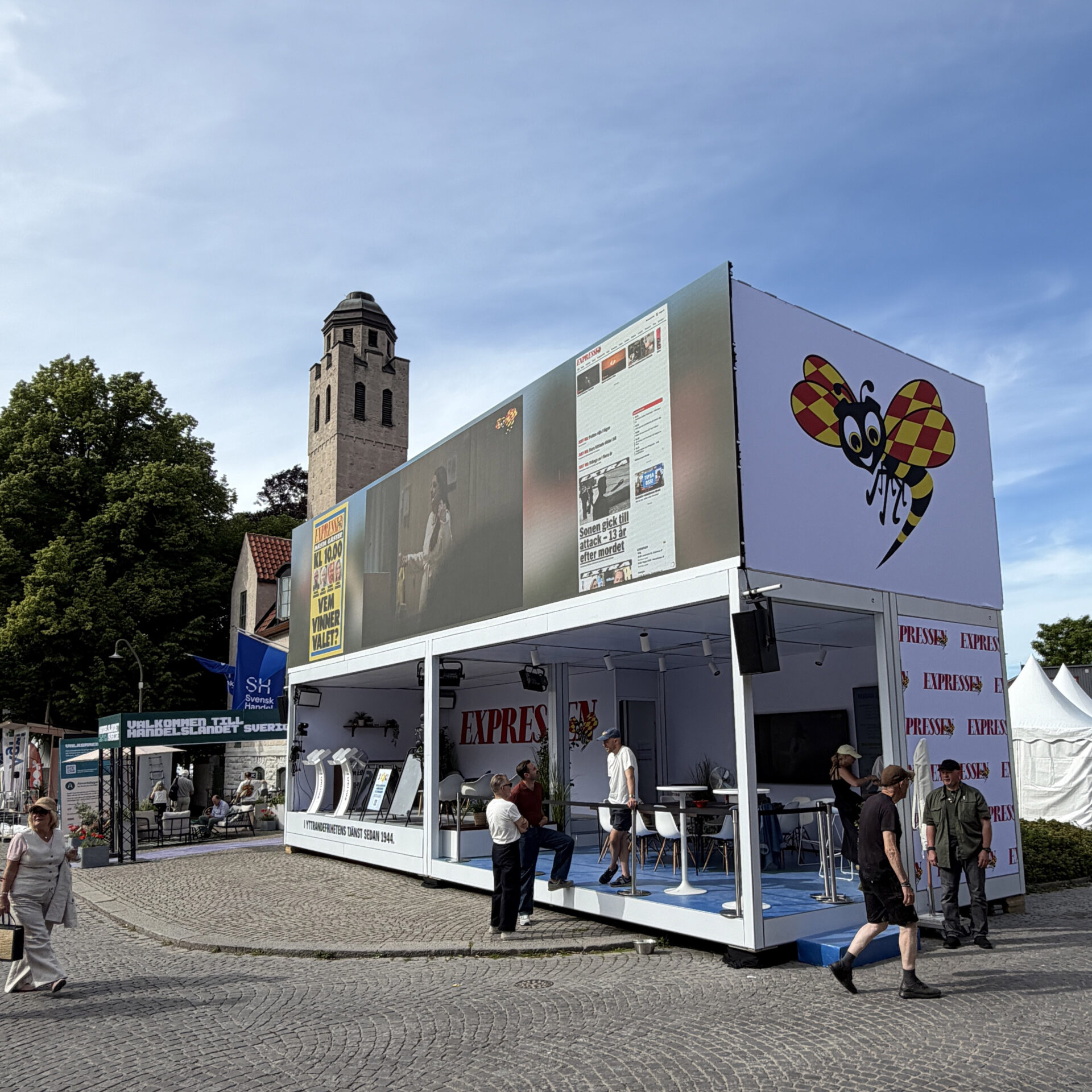 A modern white pavilion with large posters, including a cartoon bee, stands on a cobblestone square. People walk nearby, and a tall stone tower rises in the background under a blue sky.