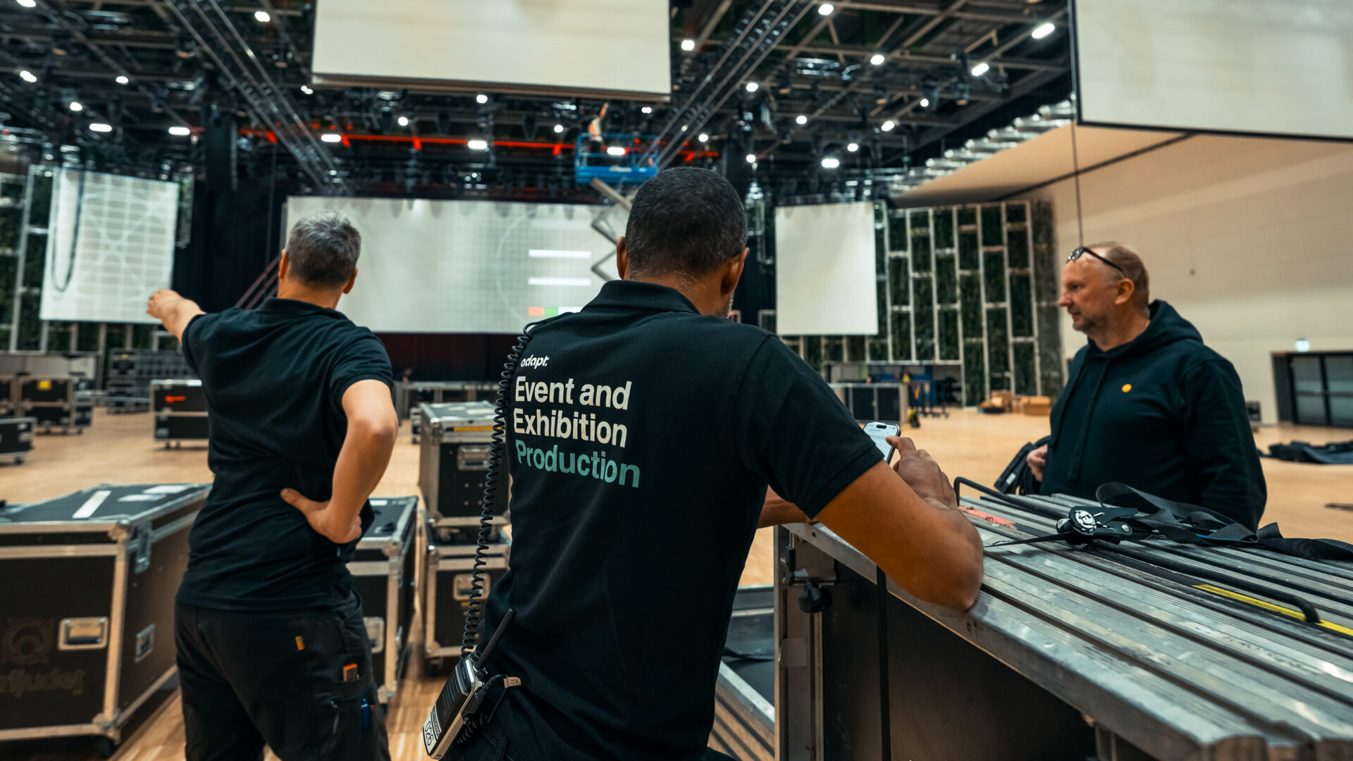 Three men wearing black shirts, two with Event and Exhibition Production written on the back, set up equipment in a large, modern event hall with hanging screens and stage lighting.