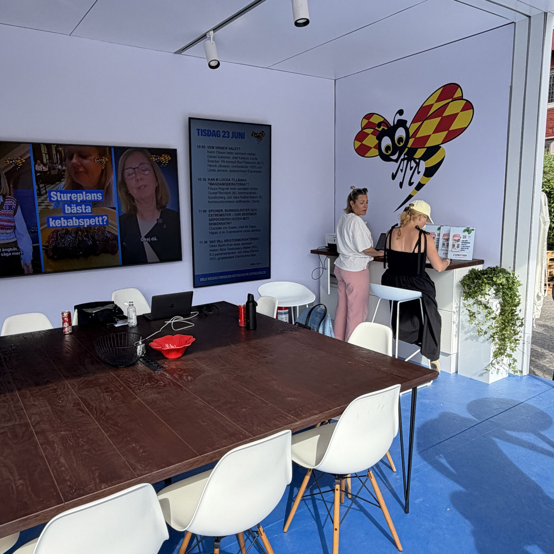 A bright room with a large wooden table surrounded by white chairs, two women standing near a counter, a large bee mural on the wall, and digital screens displaying information and photos.