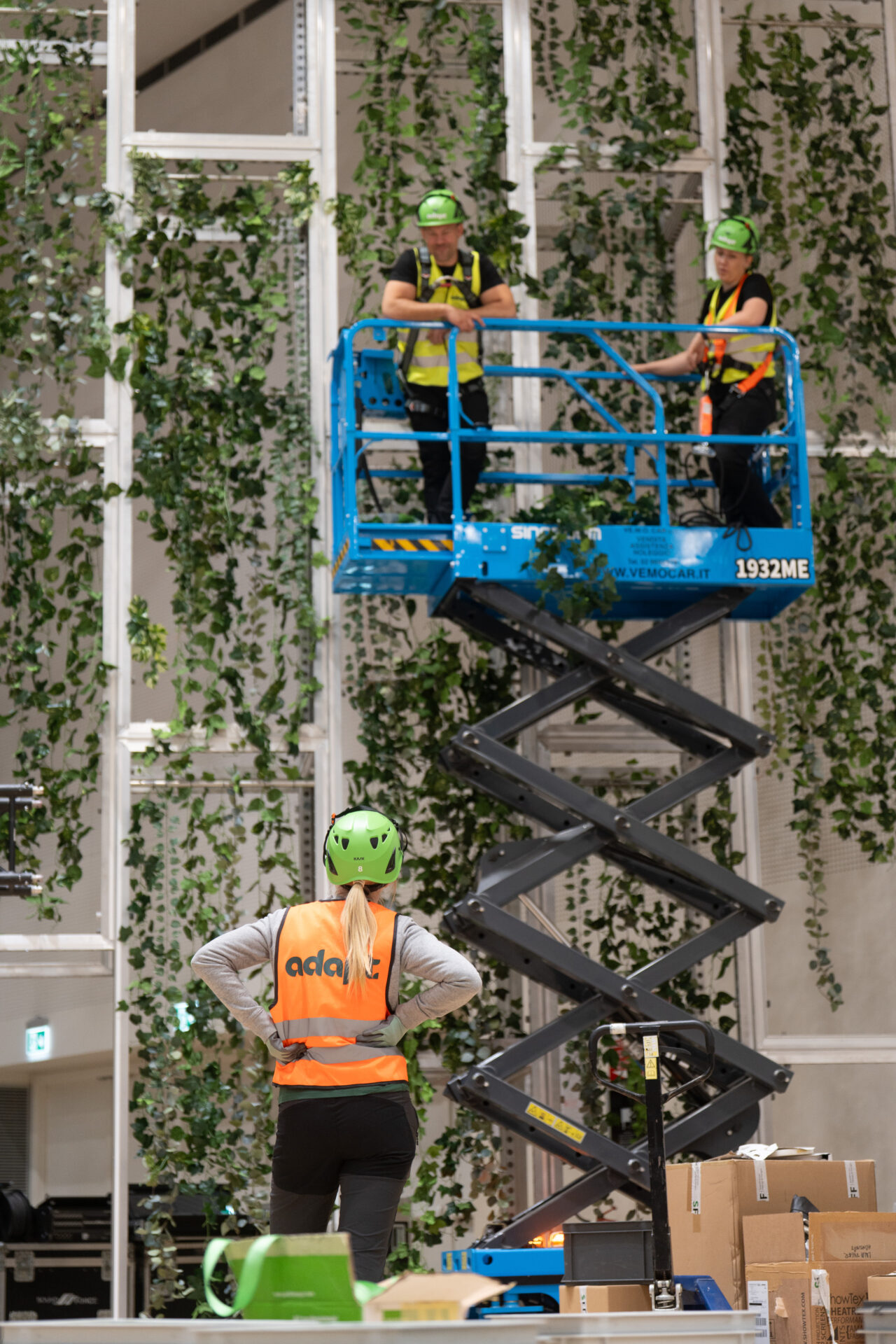 Three workers in safety gear and vests, one on the ground and two on a blue scissor lift, install or inspect greenery on a tall indoor wall. Boxes and equipment are visible in the foreground.