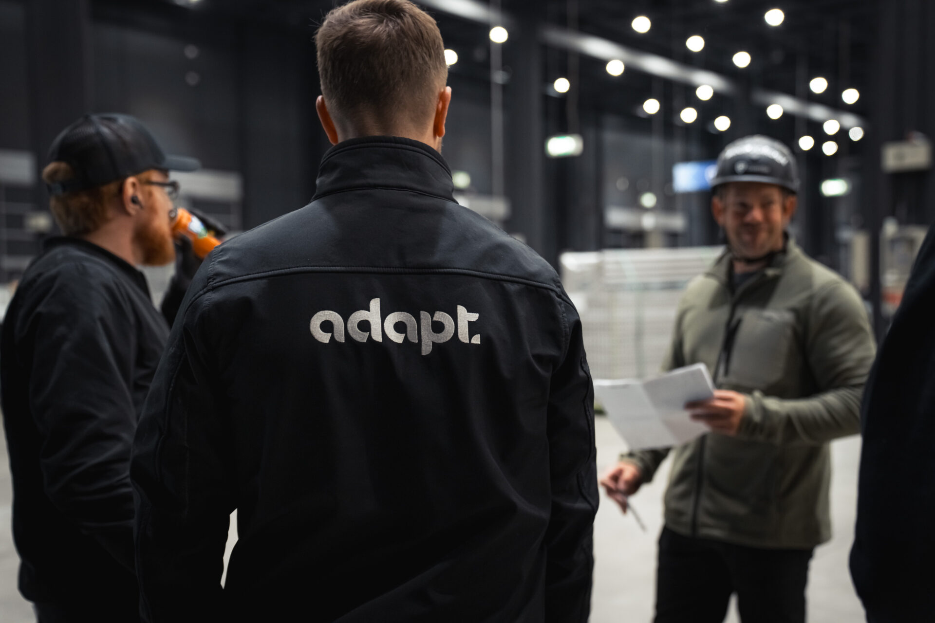 Three men stand together in a large, indoor industrial space. One mans jacket has the word adapt printed on the back. Another man is holding papers and smiling during their conversation.