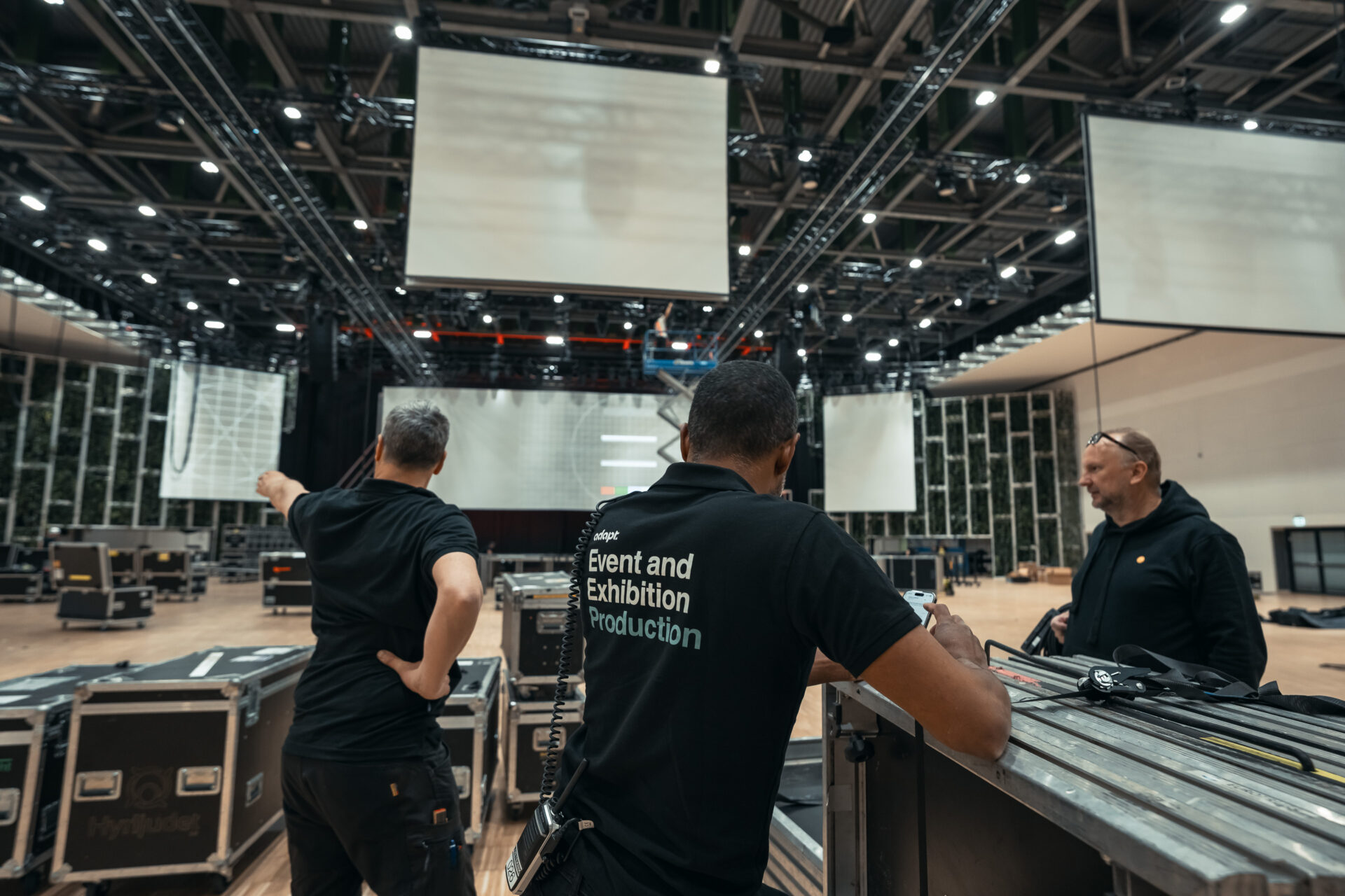 Three crew members in black shirts stand in a large indoor venue, surrounded by equipment and large screens, preparing for an event or exhibition. One person points towards the stage area.