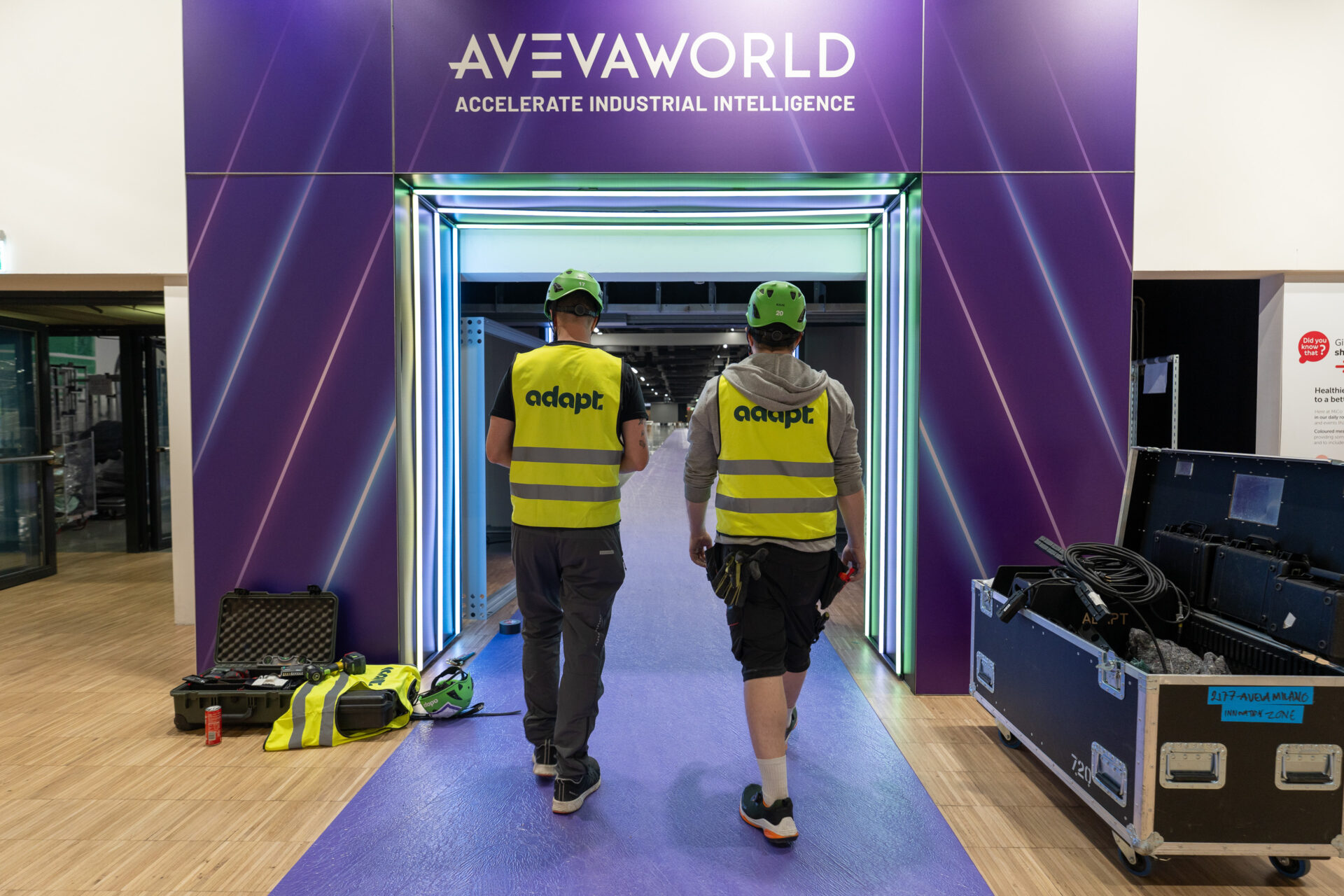 Two workers in yellow safety vests and green helmets walk through a neon-lit tunnel under a sign reading AVEVA WORLD Accelerate Industrial Intelligence, surrounded by equipment cases and tools.