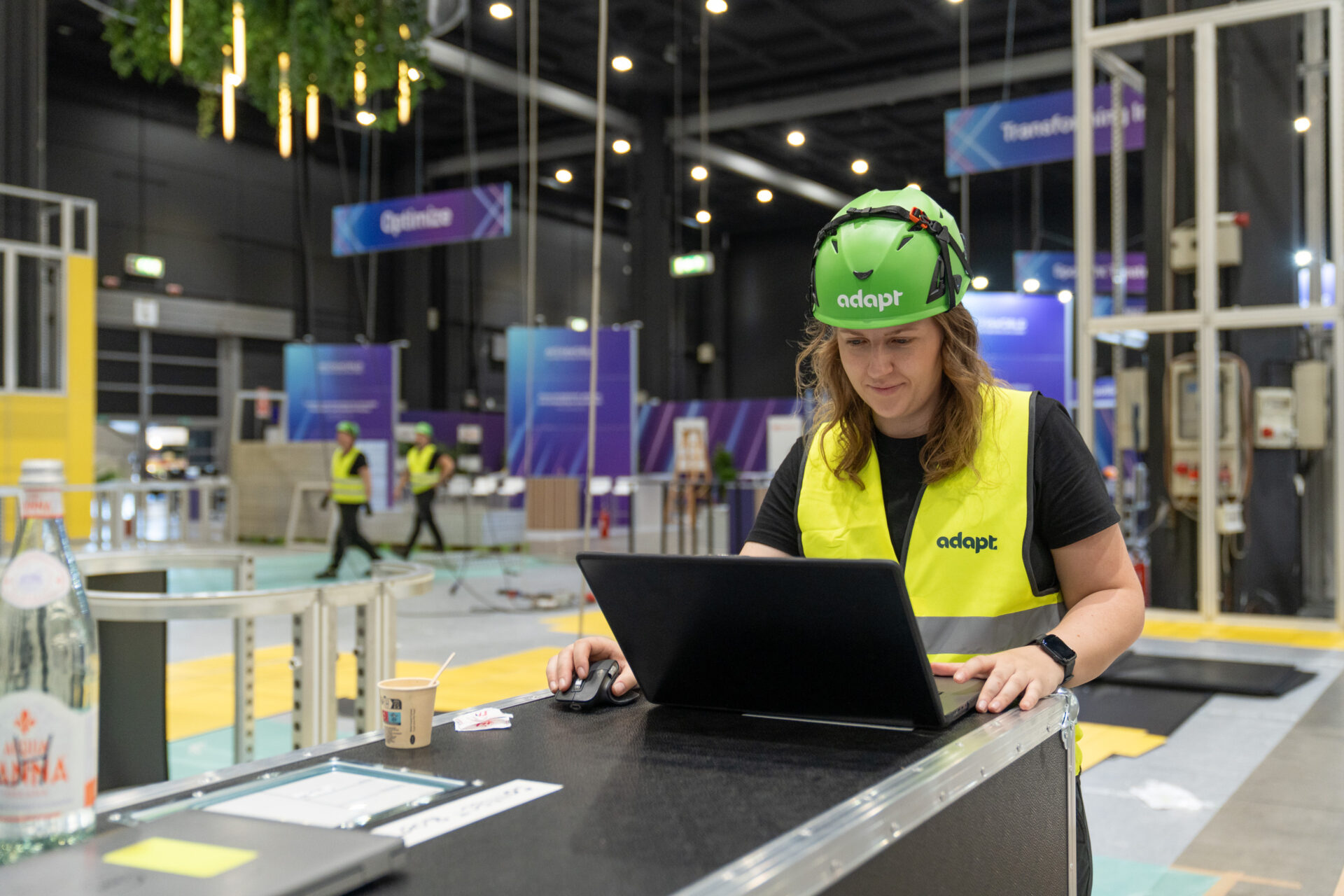 A person wearing a green helmet and yellow vest works on a laptop in an indoor event setup area with other workers and equipment in the background.