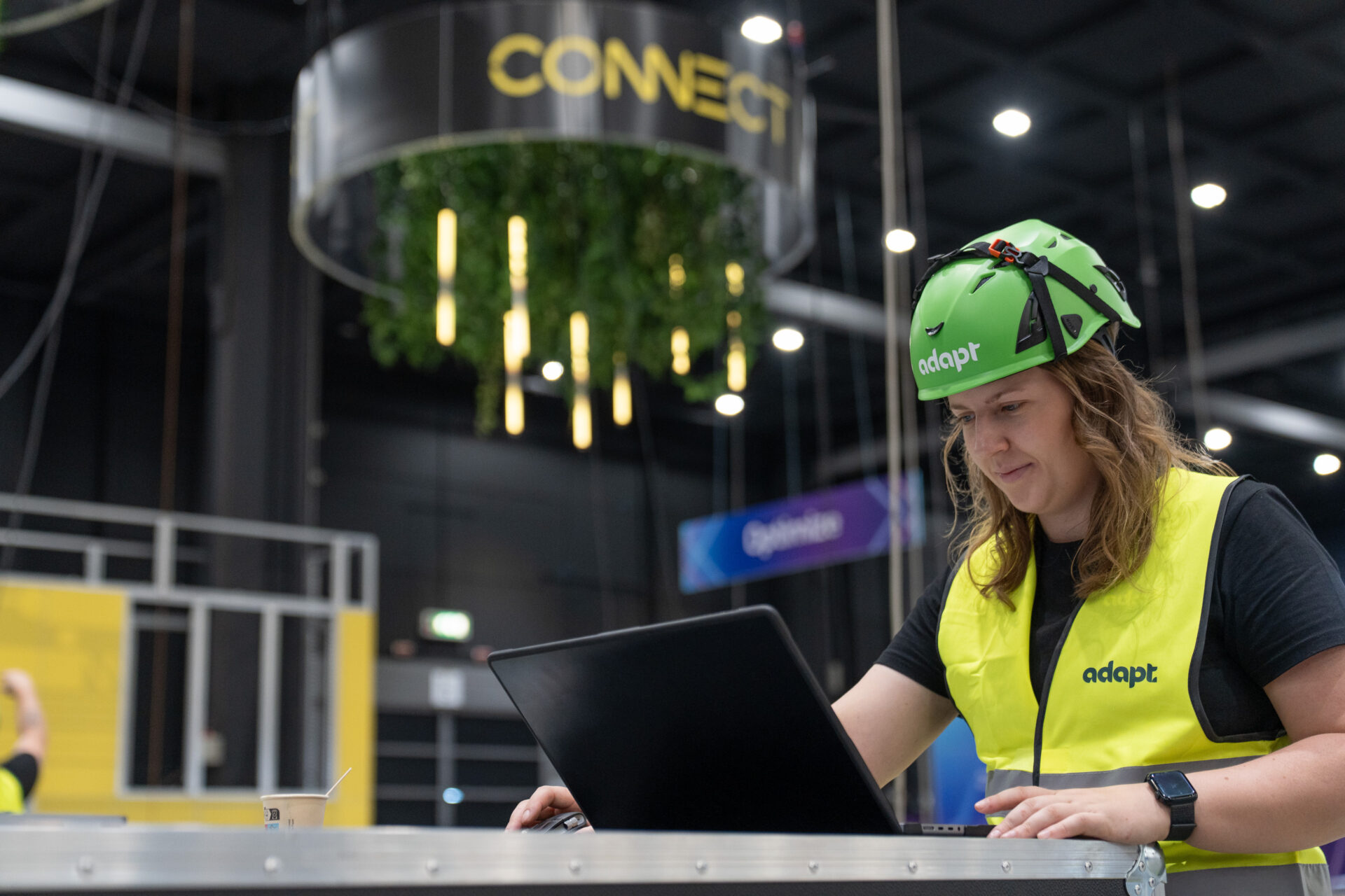 A person wearing a green helmet and yellow adapt vest works on a laptop indoors, with a CONNECT sign and hanging lights visible in the background.