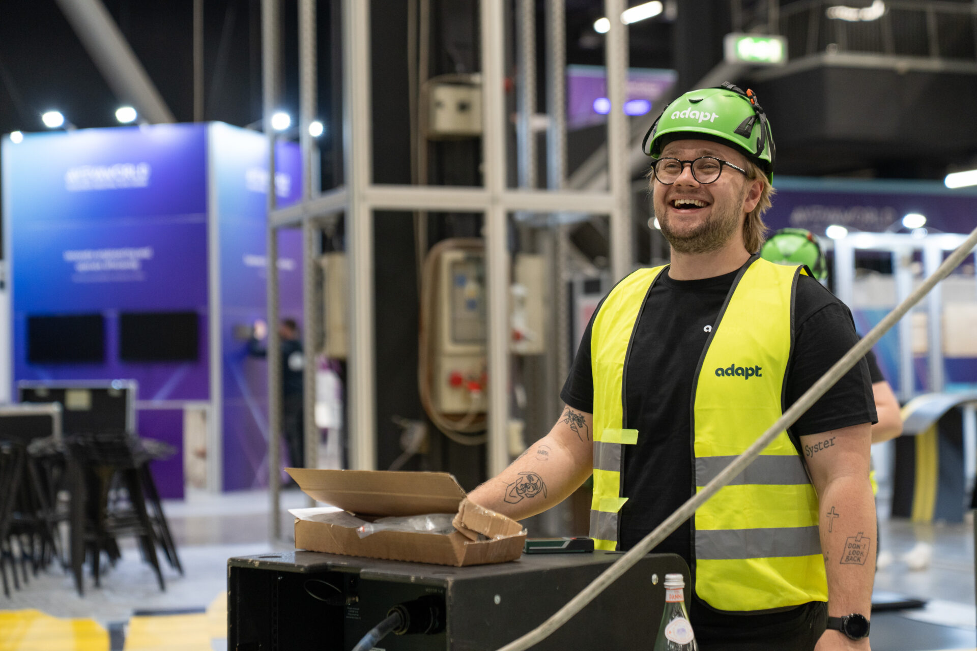 A smiling person wearing a green hard hat, glasses, and a yellow adapt safety vest stands indoors by a cart with pizza boxes, surrounded by industrial equipment and exhibition booths.