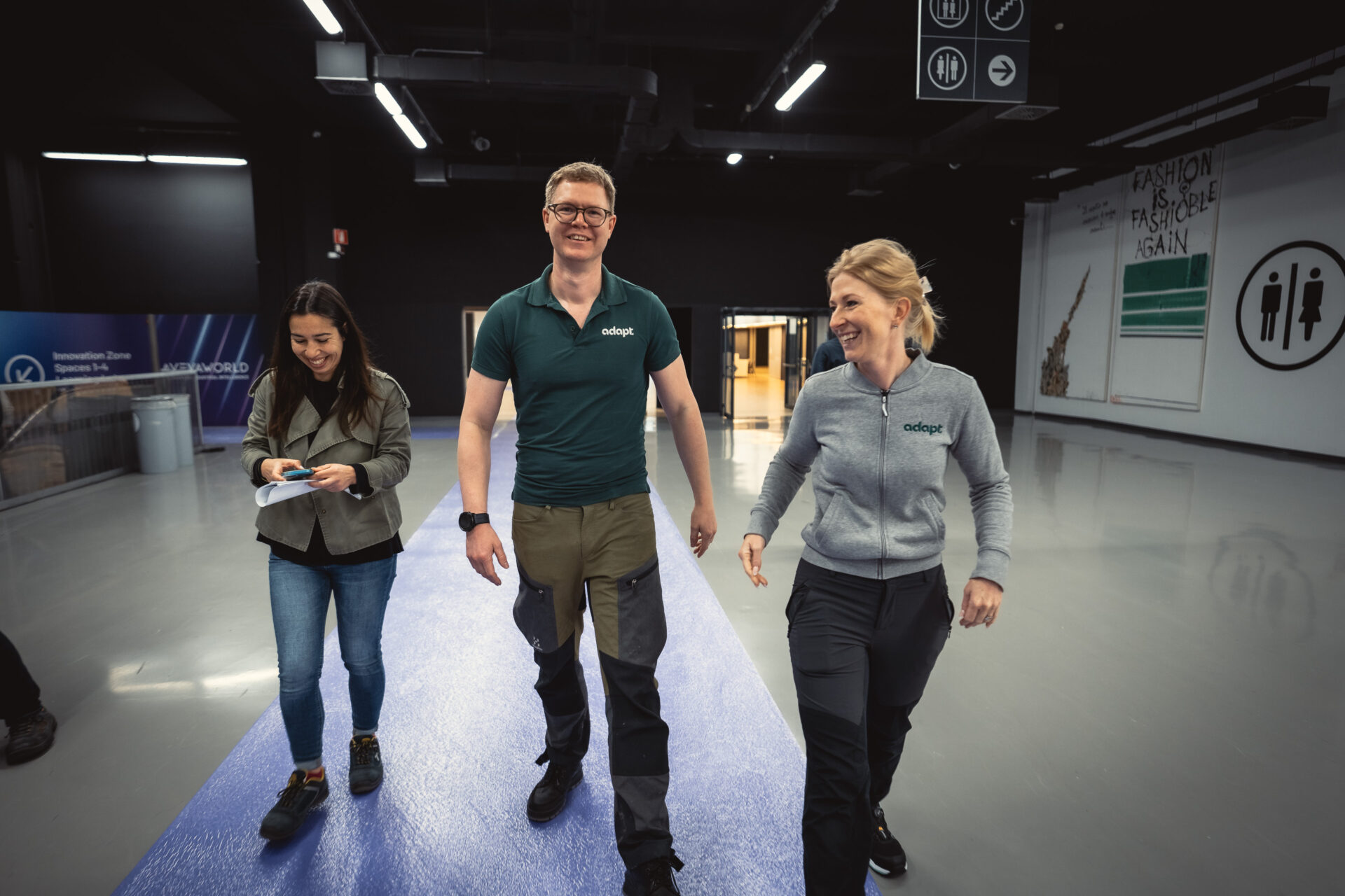 Three people, two women and one man, walk together indoors on a blue carpet. They are smiling and appear to be engaged in conversation. Signs and banners are visible on the walls in the background.