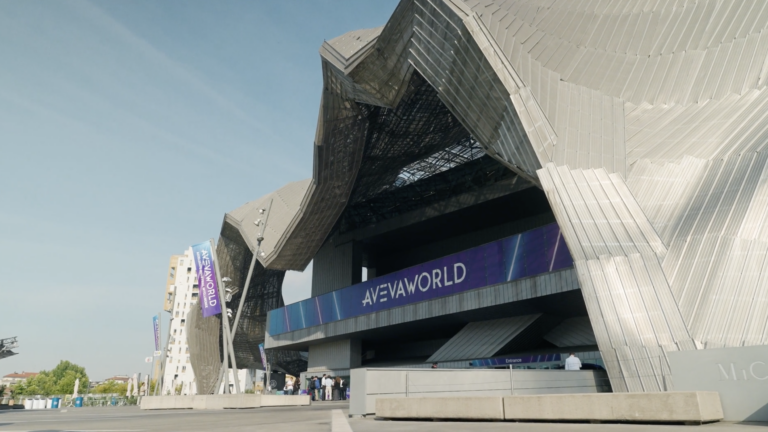 Modern building with angular, metallic architecture and a large AVEVA World sign above the entrance. People are gathered near the entrance, and blue banners with AVEVA WORLD are displayed outside.