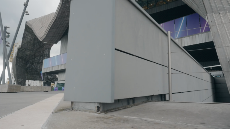 A modern building with large geometric concrete structures and purple glass panels, seen from a low angle on a cloudy day. The scene includes a sidewalk and angled architectural elements.