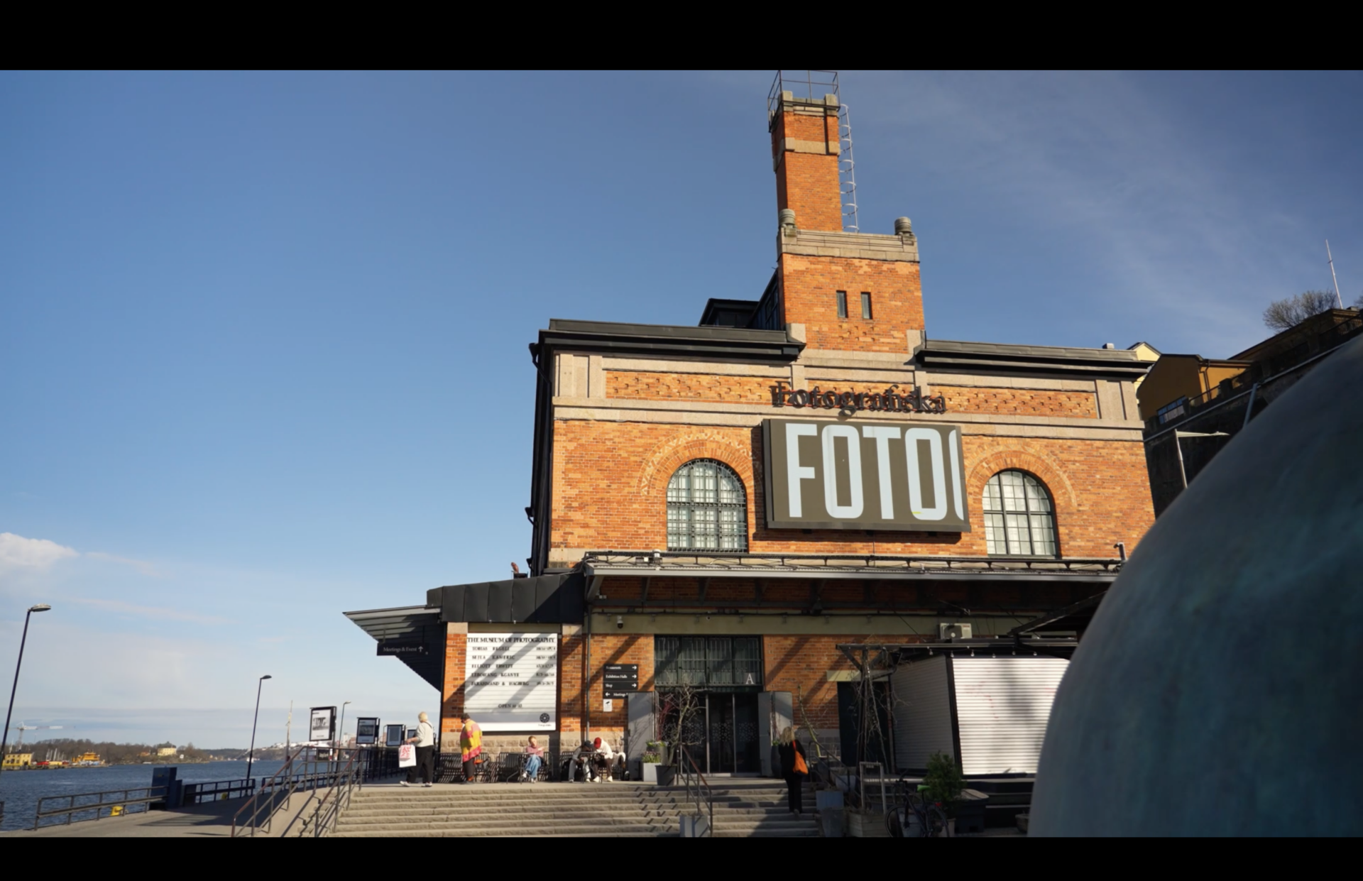 A large brick building with a sign reading FOTO above the entrance, situated by the waterfront under a clear blue sky, with people gathered near the steps outside.