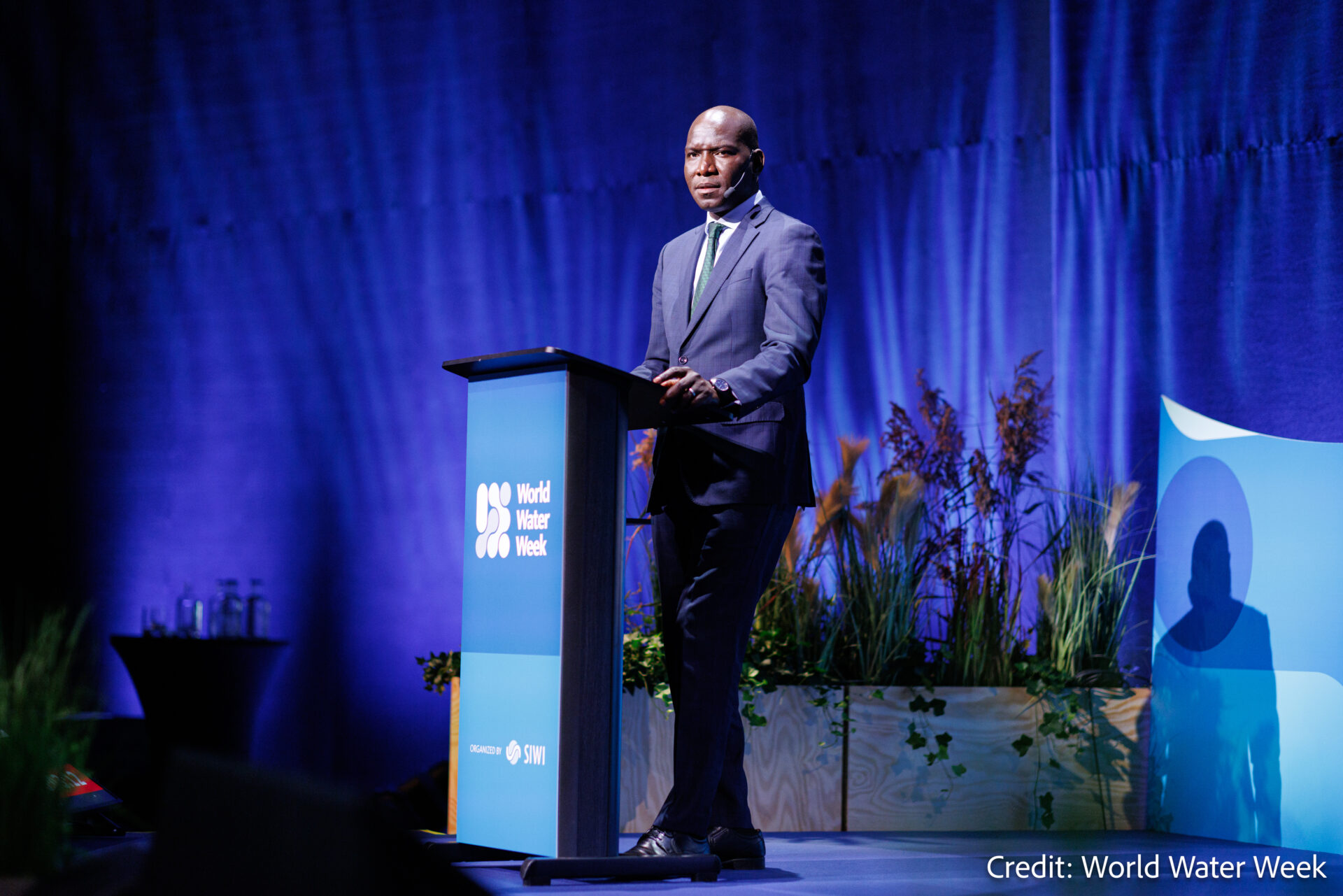 A man in a suit speaks at a blue podium labeled World Water Week, with plants and a blue backdrop behind him. The photo is credited to World Water Week.