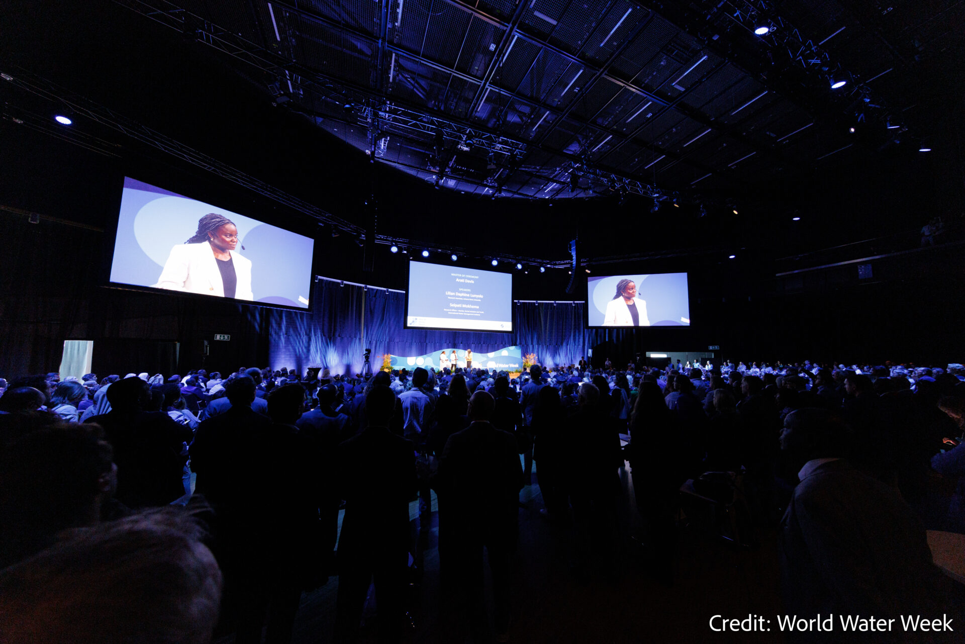 A large audience watches a speaker on stage at a conference. Two large screens display the speaker and presentation slides. The room is dimly lit. Credit: World Water Week.