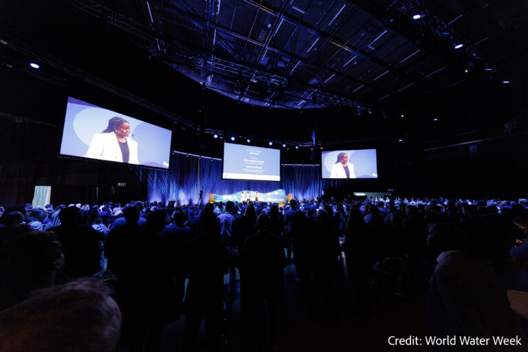 A large audience watches a speaker on stage at a conference. Two large screens display the speaker and presentation slides. The room is dimly lit. Credit: World Water Week.