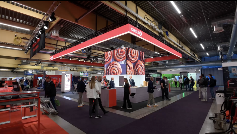 An indoor trade show features a booth with a red CIRCO logo and images of pastries above. People walk and interact around the booth, with various other exhibits and machines visible in the background.