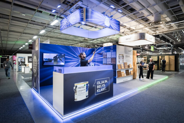 A modern trade show booth with bright lighting showcases Osmo brand products. A person stands behind the counter, while others interact nearby. Displays feature cans, wooden samples, and informational signage under an industrial ceiling.