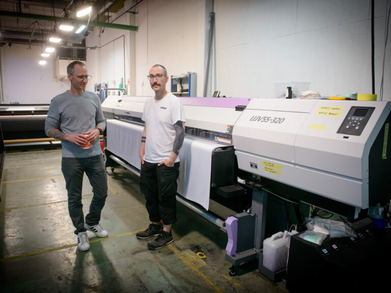 Two men stand next to a large UV printer in a brightly lit industrial workspace; one holds a red cup while the other has his hand in his pocket.