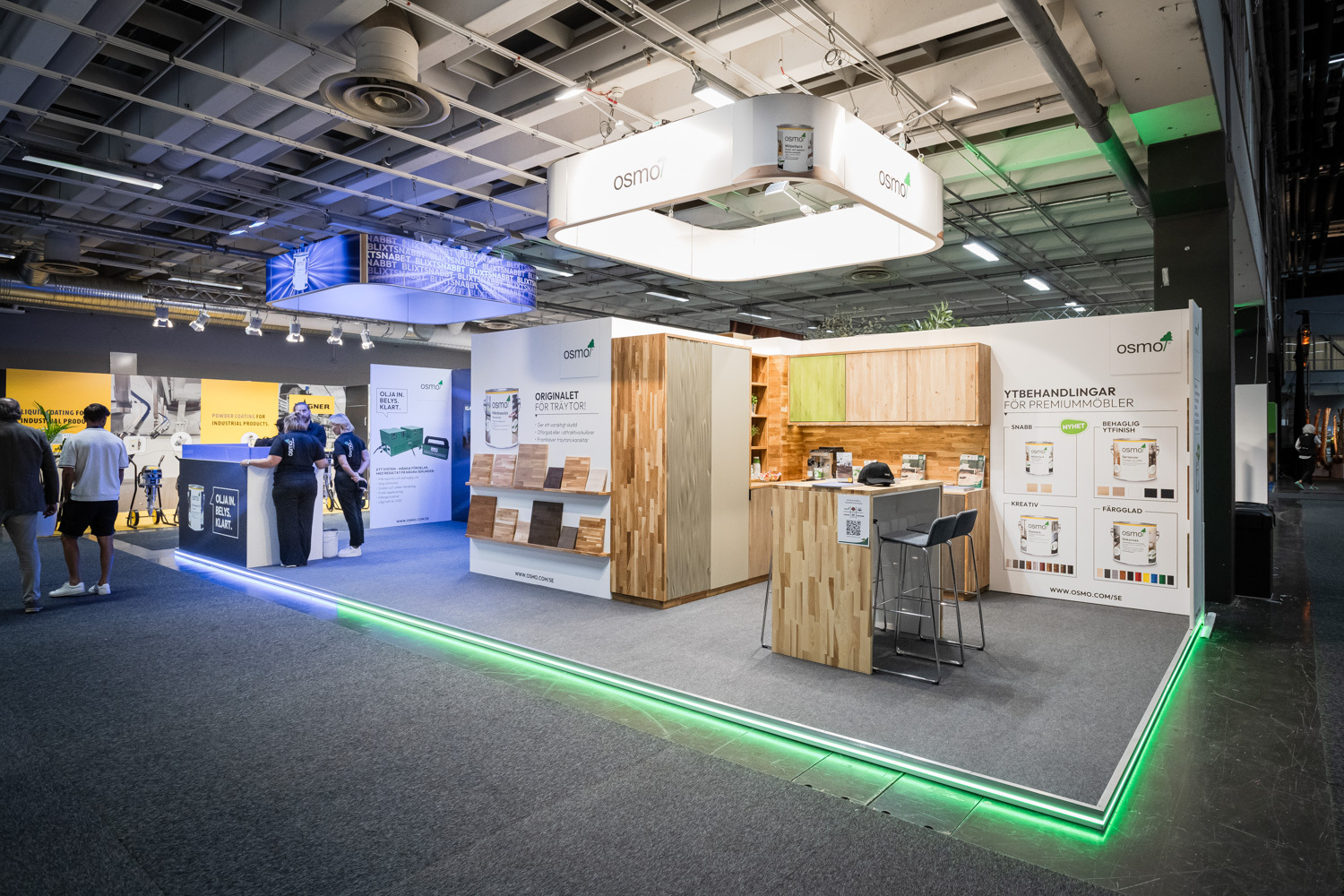 A modern exhibition booth with wood samples, product displays, and informational posters. Several people are walking nearby in a large, well-lit convention hall.