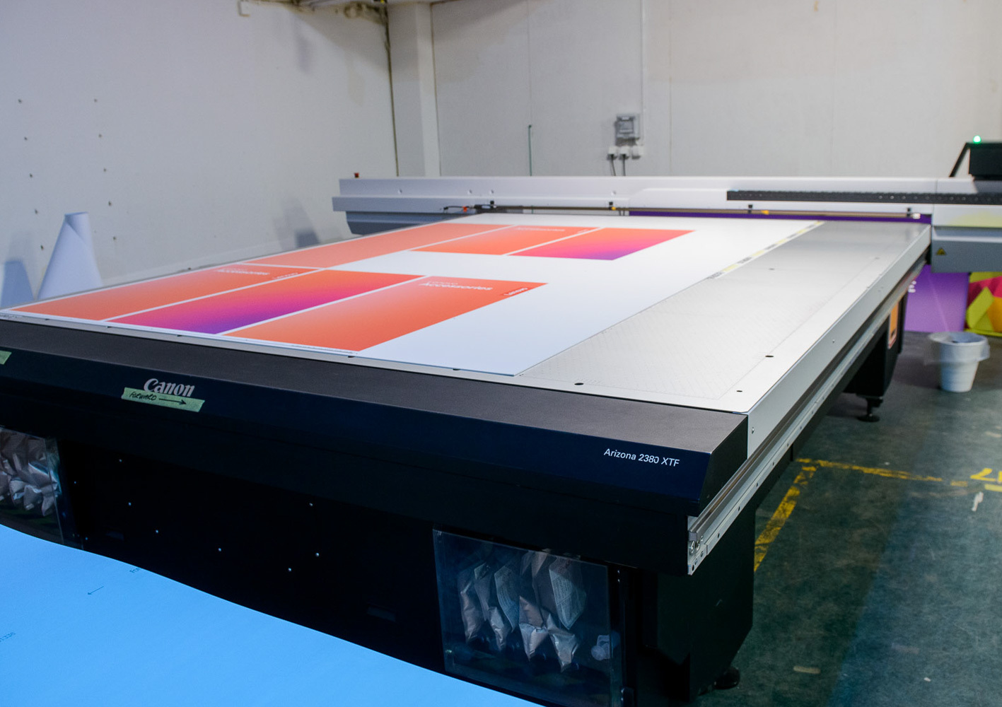 A large-format Canon Arizona 2260 XTF printer prints colorful posters on a flatbed inside an industrial workspace.