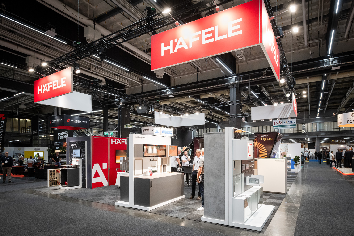A modern trade show booth for Häfele features sleek displays, lighting, and signage under bright overhead lights, with visitors walking and exploring the exhibits in a spacious indoor venue.