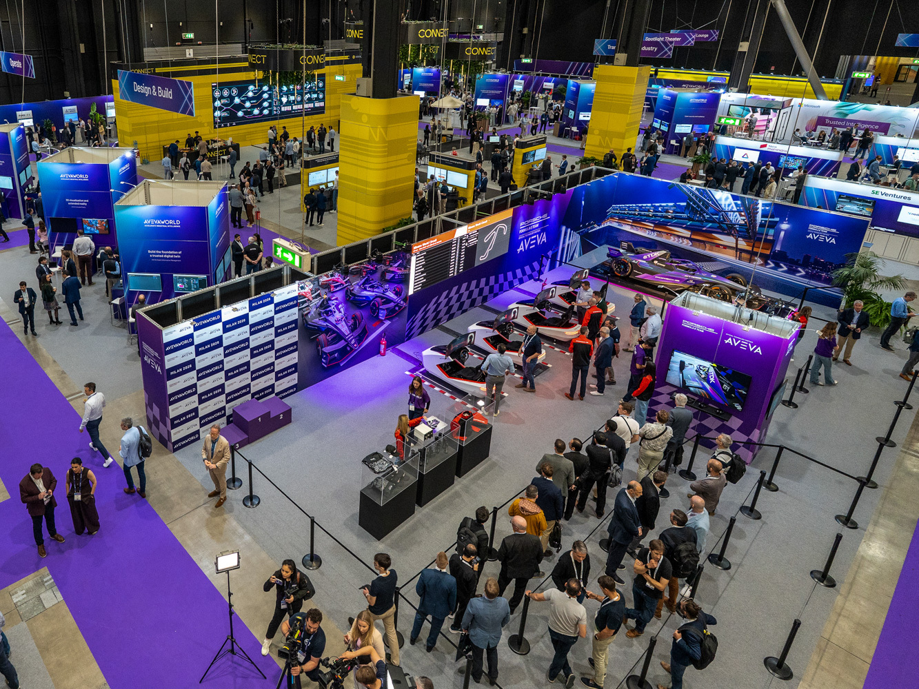 A busy tech expo hall with colorful booths, racing car displays, and crowds of people exploring exhibits and talking in groups. Wide walkways and banners are visible throughout the venue.