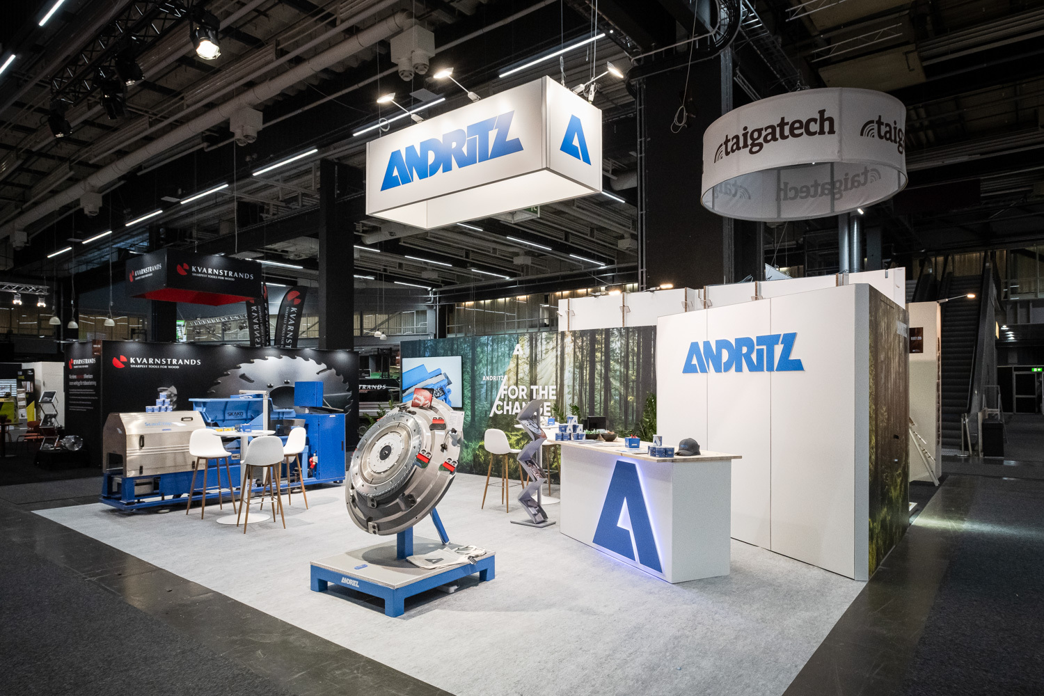 ANDRITZ trade show booth with machinery display, branded signs, a counter with chairs, and forest graphics in the background inside a large, well-lit exhibition hall.