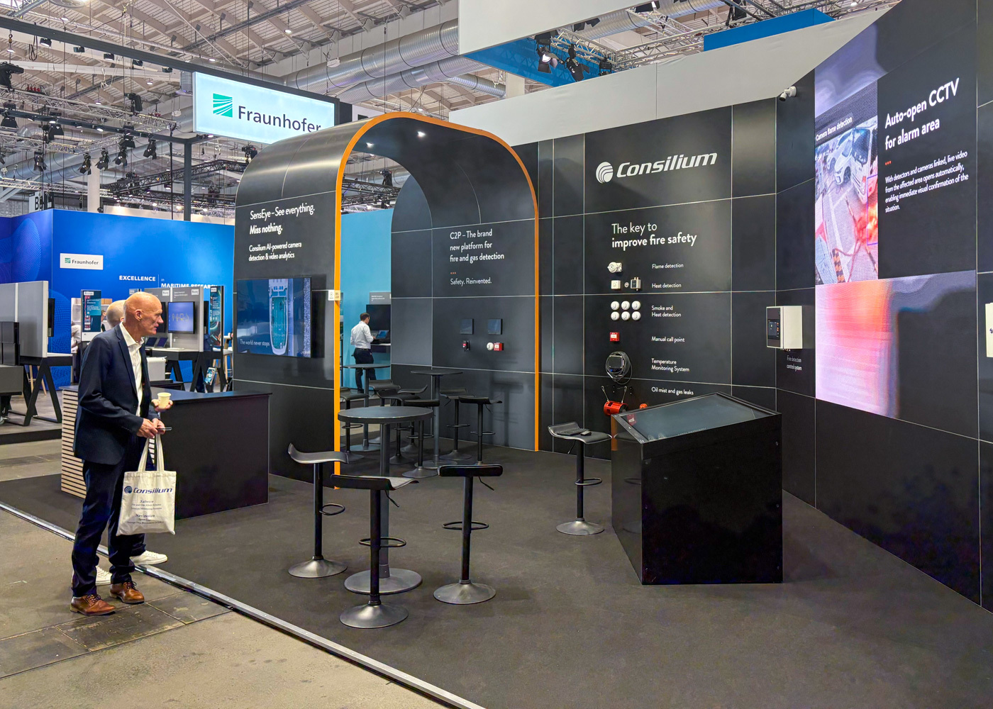 A man stands near a modern trade show booth for Consilium, featuring displays about fire safety and CCTV. The booth has barstools, a table, information panels, and technology equipment on display.