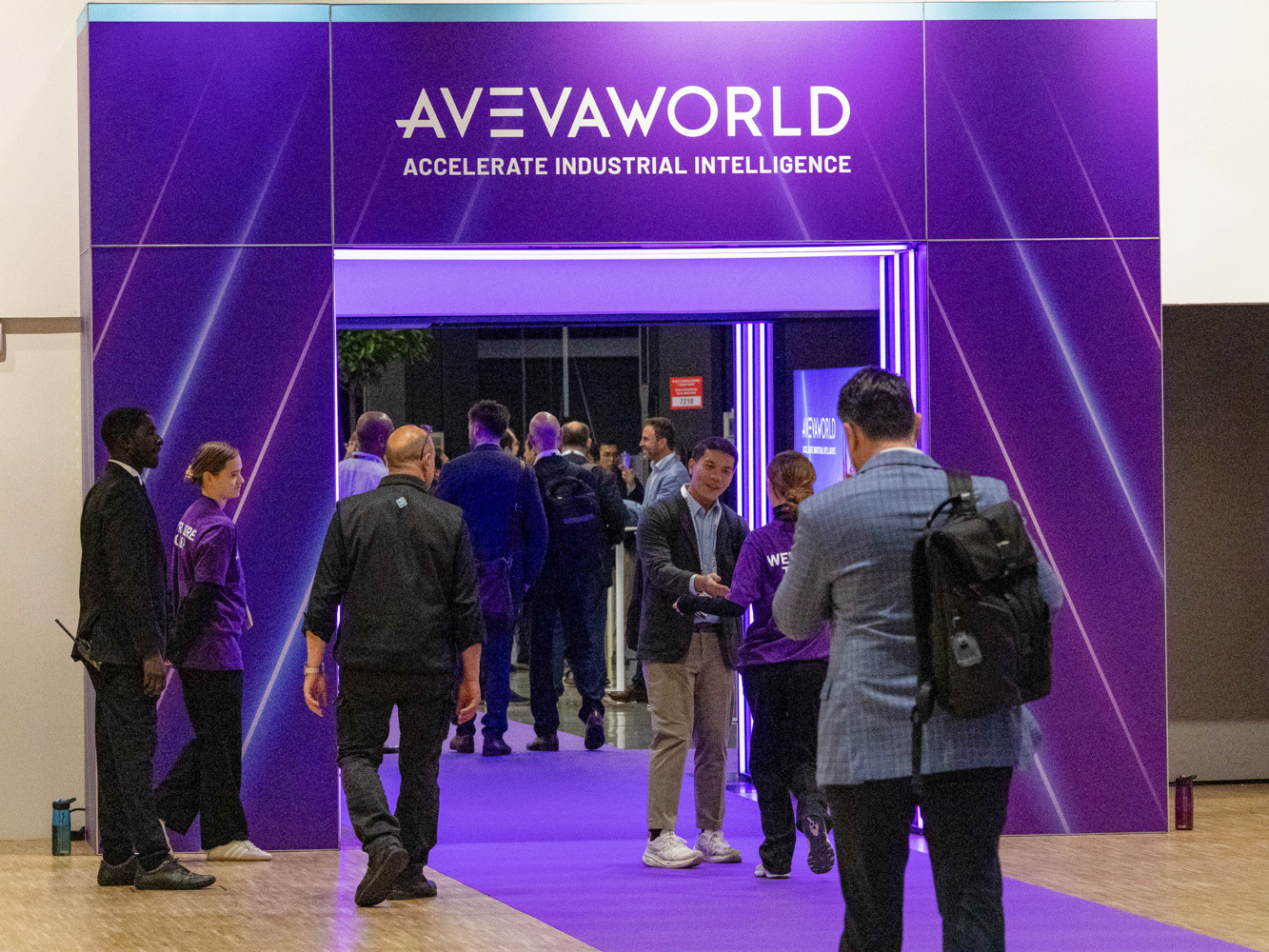 People walk through a purple entrance labeled AVEVA World Accelerate Industrial Intelligence at a conference or event. Some wear matching jackets, and one person is holding a phone.
