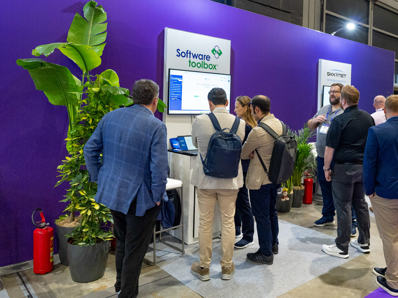 Several people gather around a Software Toolbox booth at a tech event, looking at a screen. There are potted plants and a fire extinguisher nearby, with a purple backdrop and another booth seen in the background.