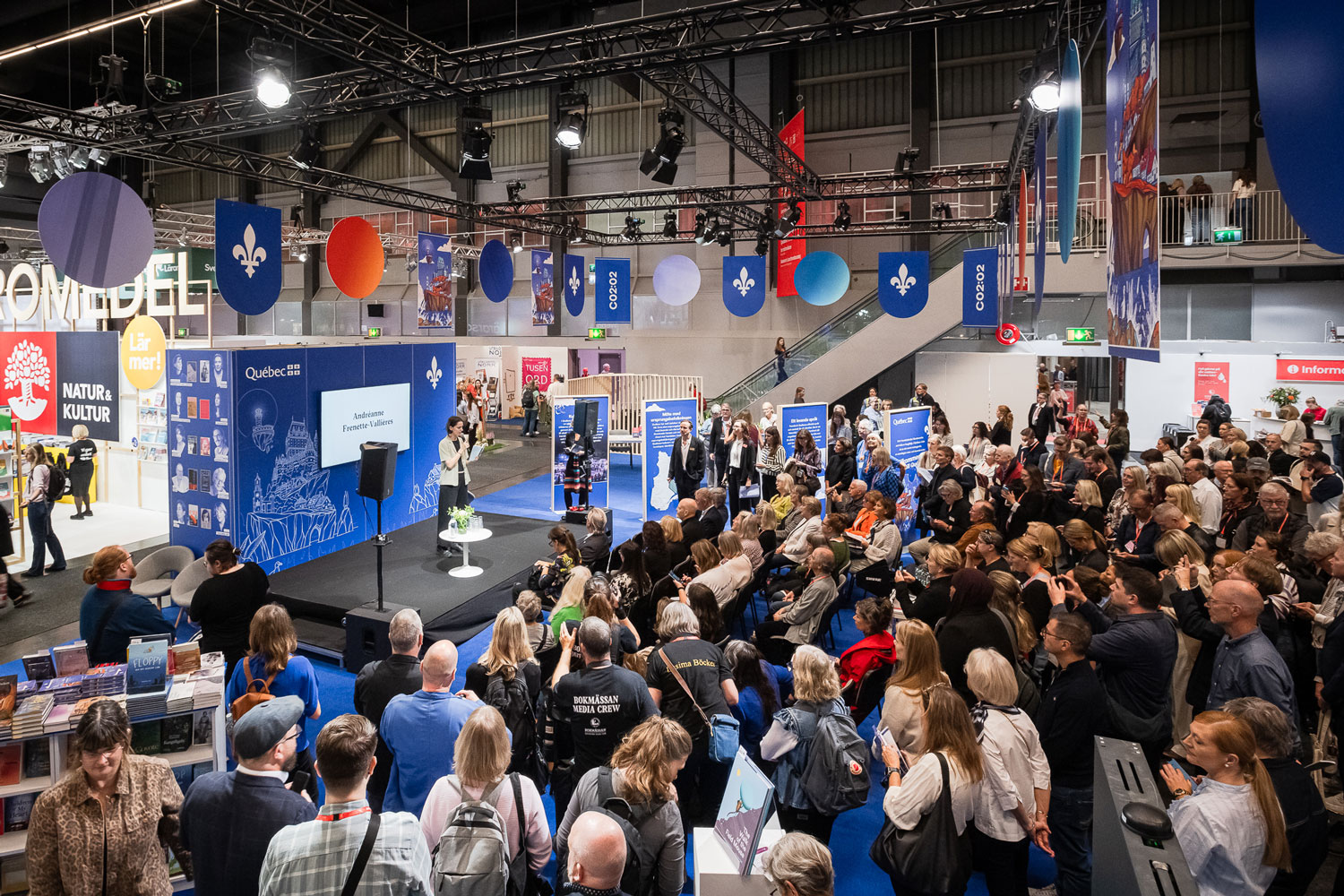 A large crowd gathers at a convention or expo, listening to a speaker on stage. Booths, banners, and books are visible, with blue Quebec flags hanging above. The setting appears lively and well-attended.
