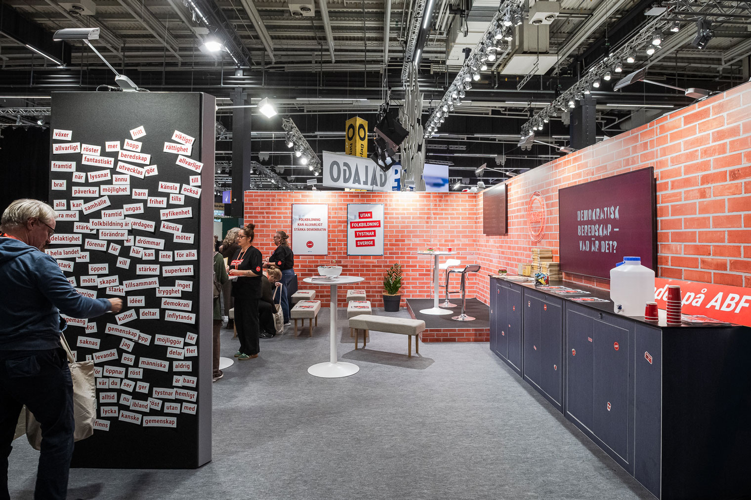A booth at a convention features brick-patterned walls, a blackboard with words, tables, chairs, and people interacting. A wall is covered with word magnets, and a counter holds various items.